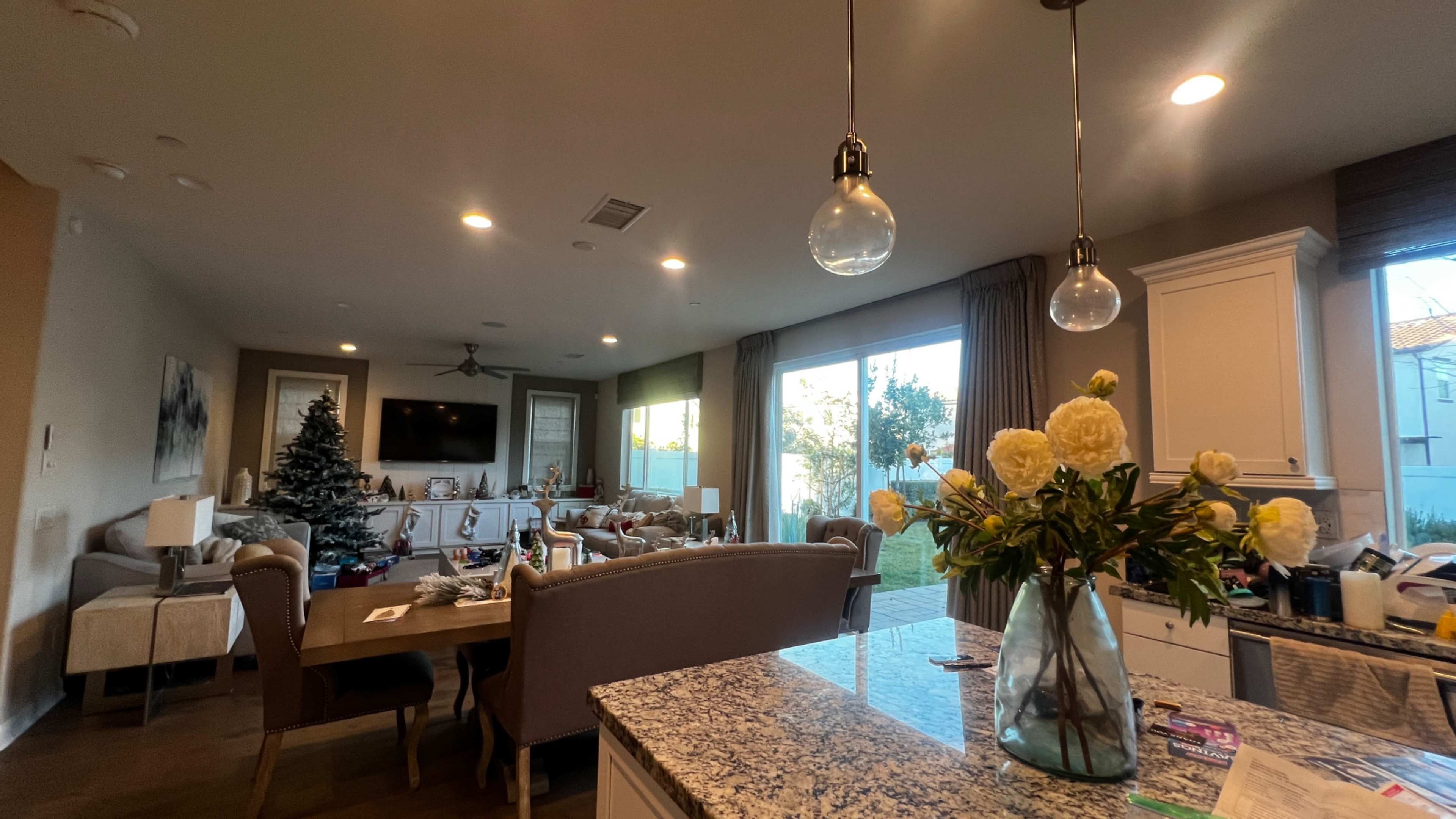 A modern living room and dining area with a Christmas tree, large windows, and a kitchen counter featuring a vase of flowers.