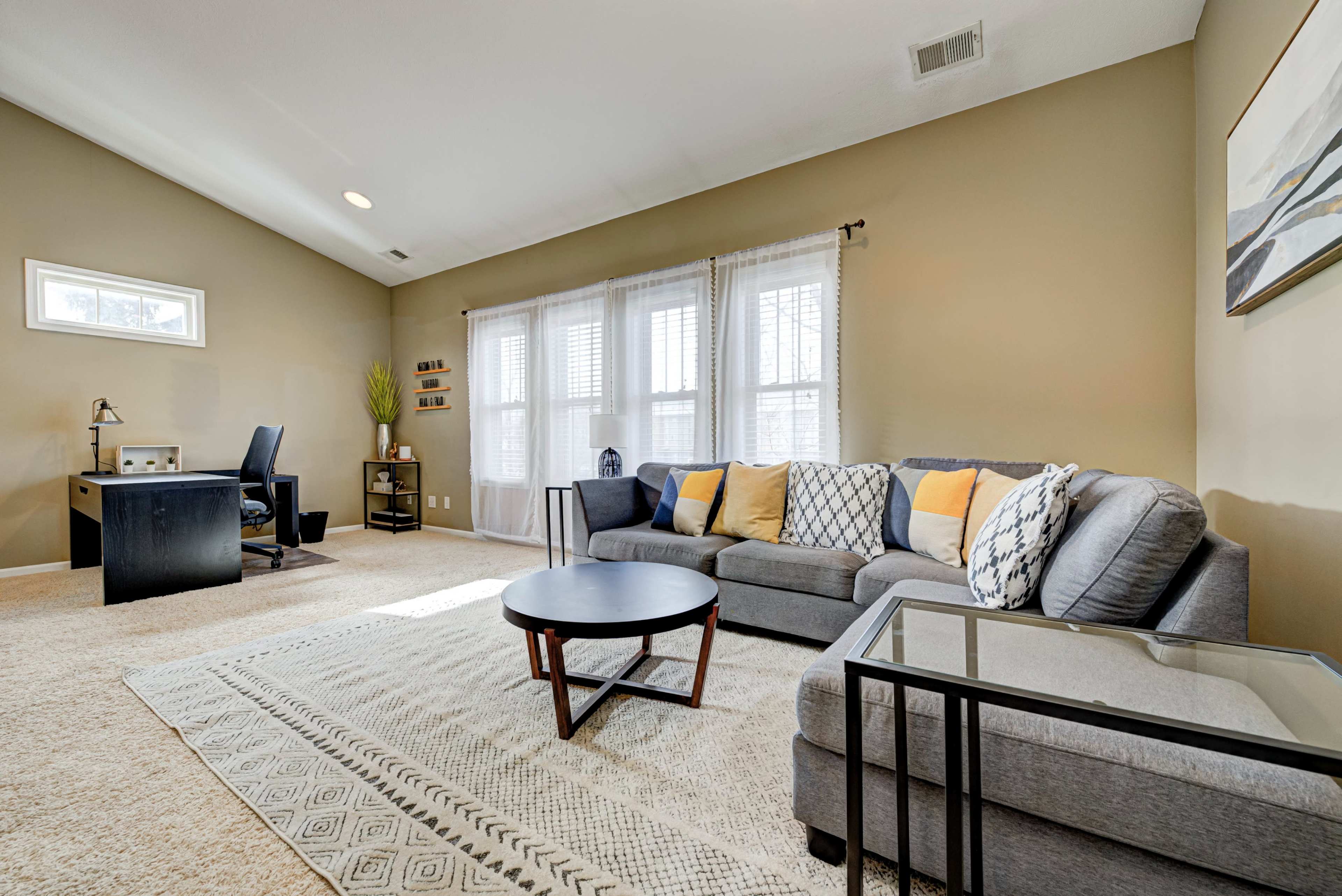 A cozy living room features a gray sofa, a round coffee table, a desk with a chair, and large windows allowing natural light to fill the space.