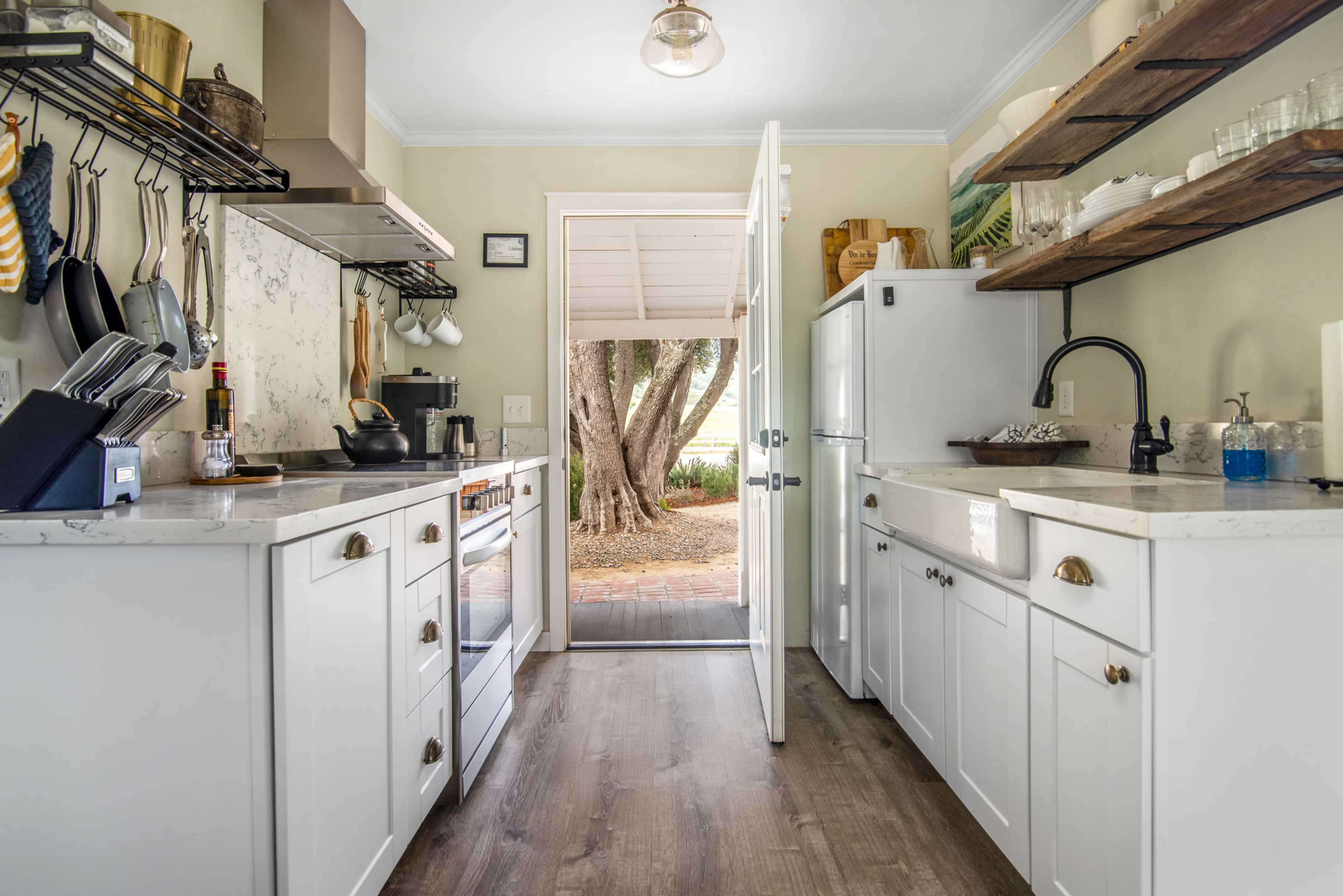 A kitchen with white cabinetry opens to an outdoor area through a door, featuring a side-by-side refrigerator and shelves with glassware.