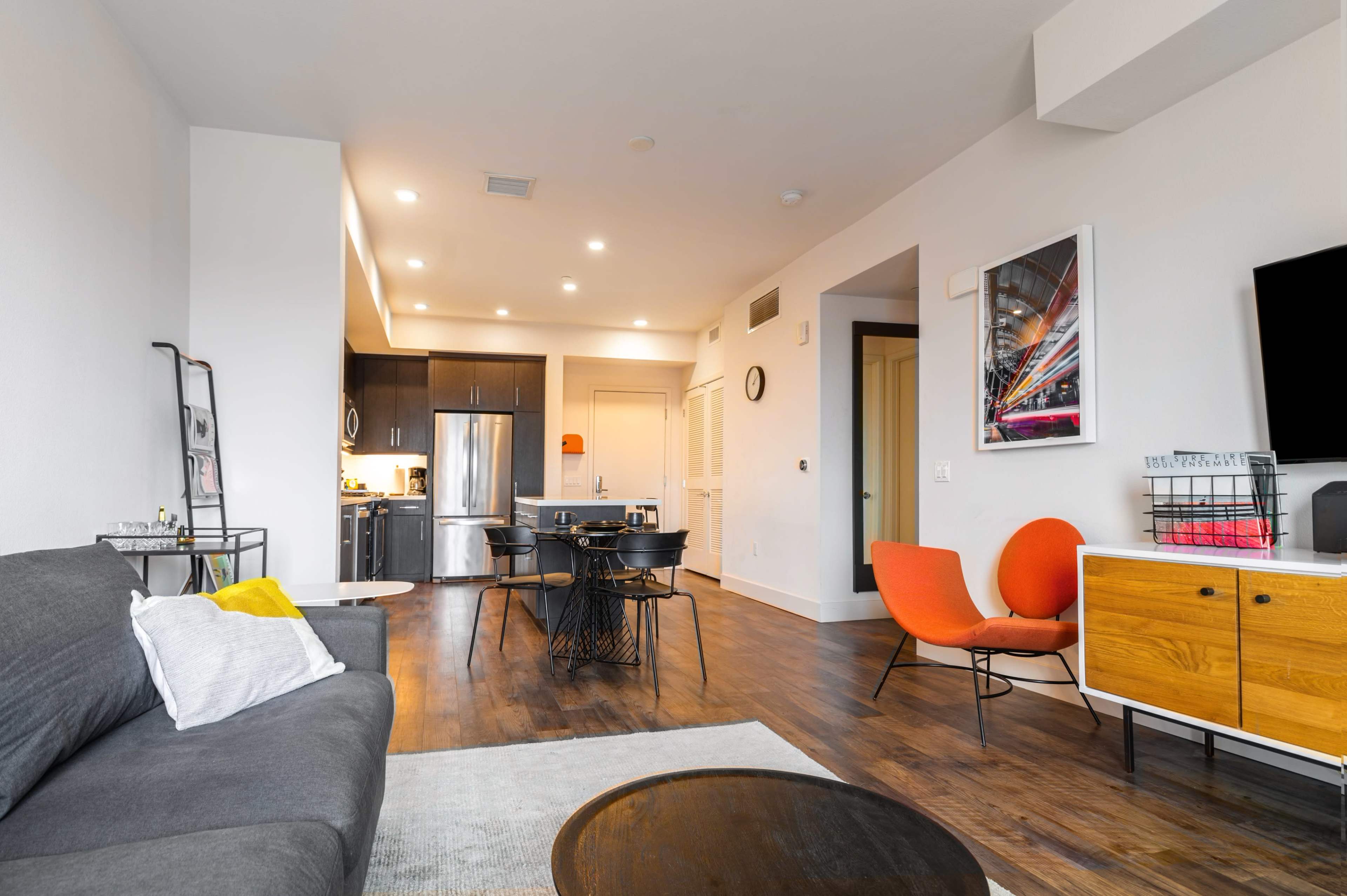A modern living area with a gray sofa, orange chairs, a round table, and an open kitchen with dark cabinets.