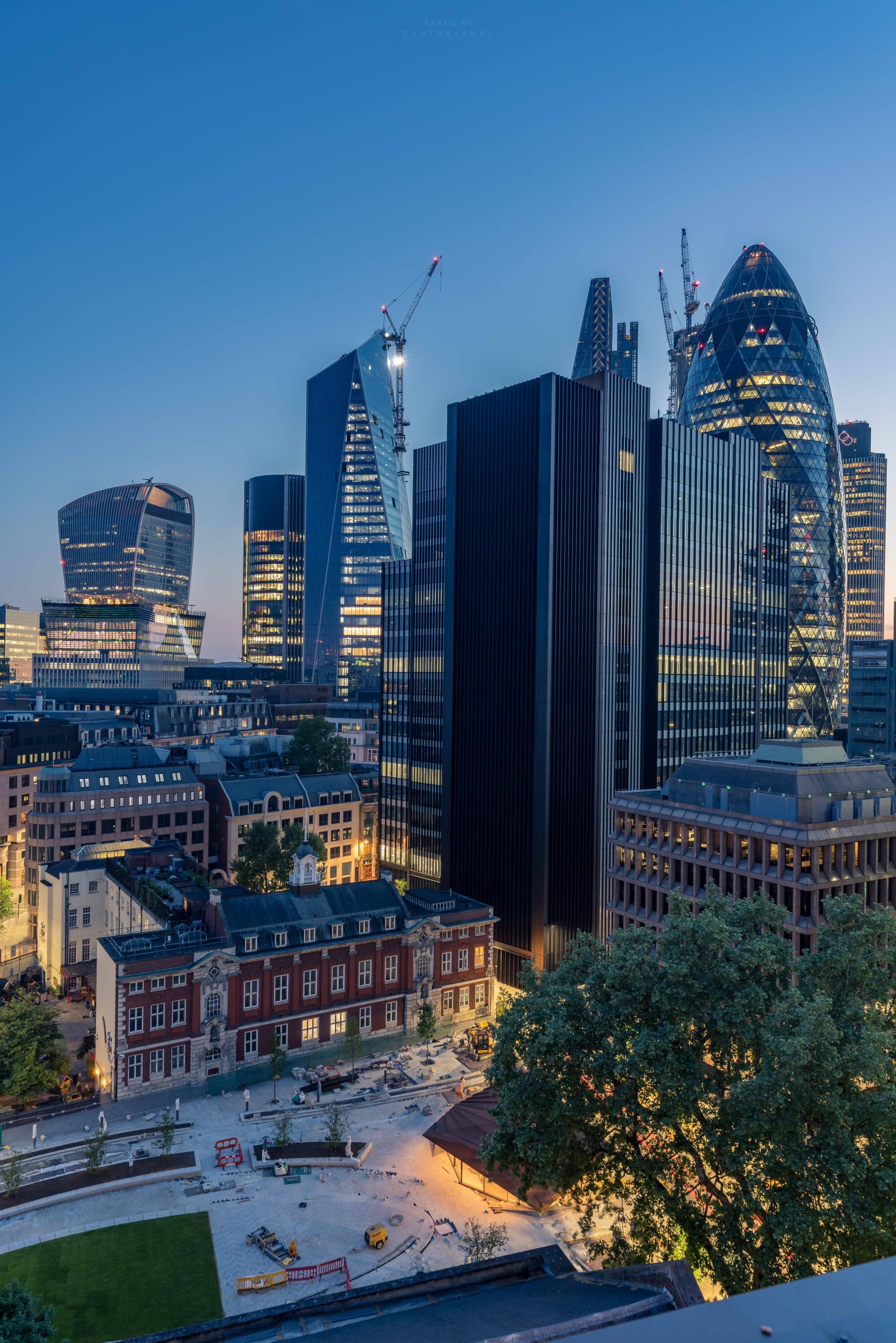 The image shows a city skyline at dusk featuring a mix of modern skyscrapers and historical buildings, with construction cranes visible on some of the towers.