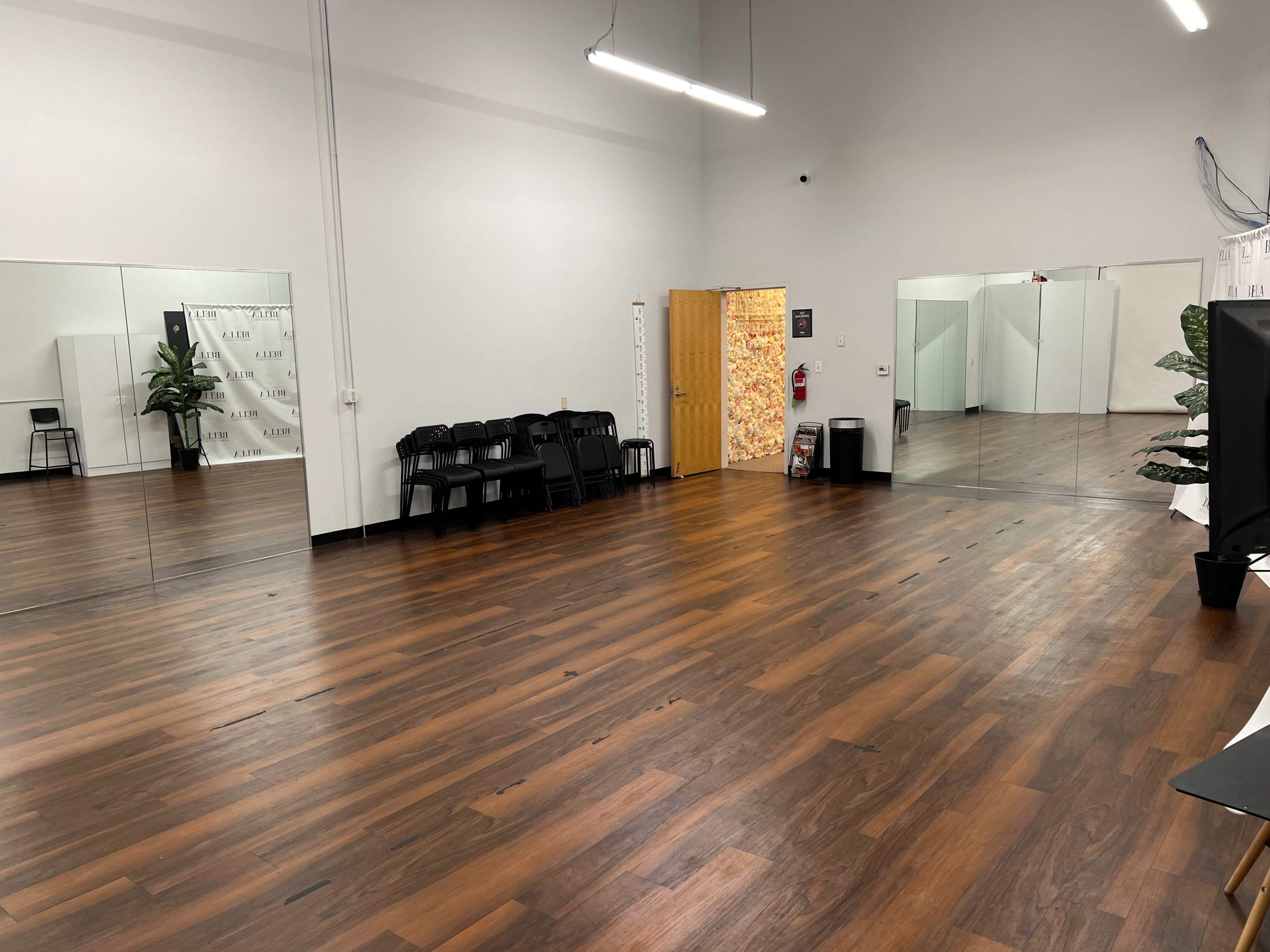 A spacious studio with wooden flooring, large mirrors on one wall, a few black chairs stacked against another wall, and a colorful door at the far end.
