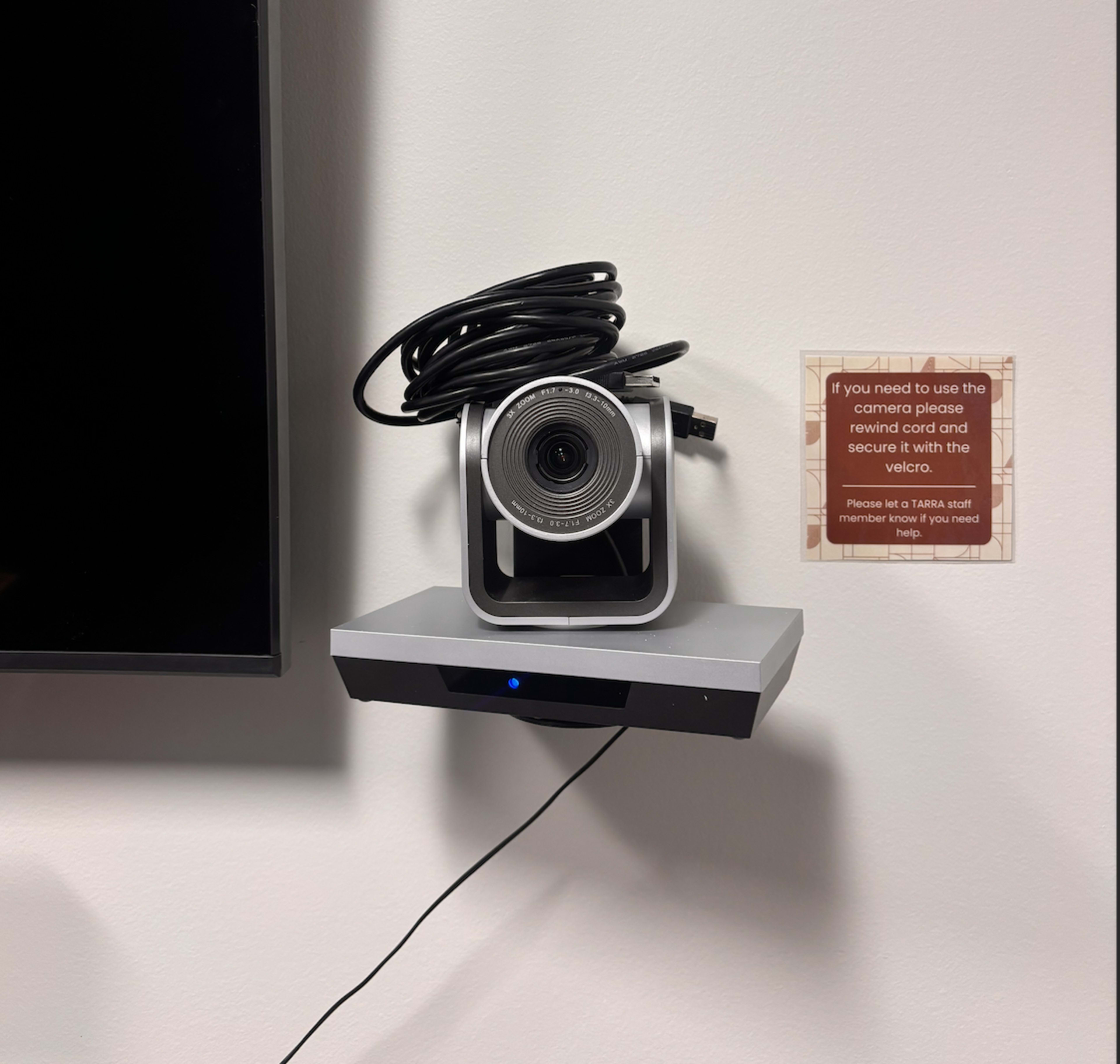 A camera with a coiled cable is mounted on a wall above a gray shelf, next to a notice instructing users to seek assistance if needed.