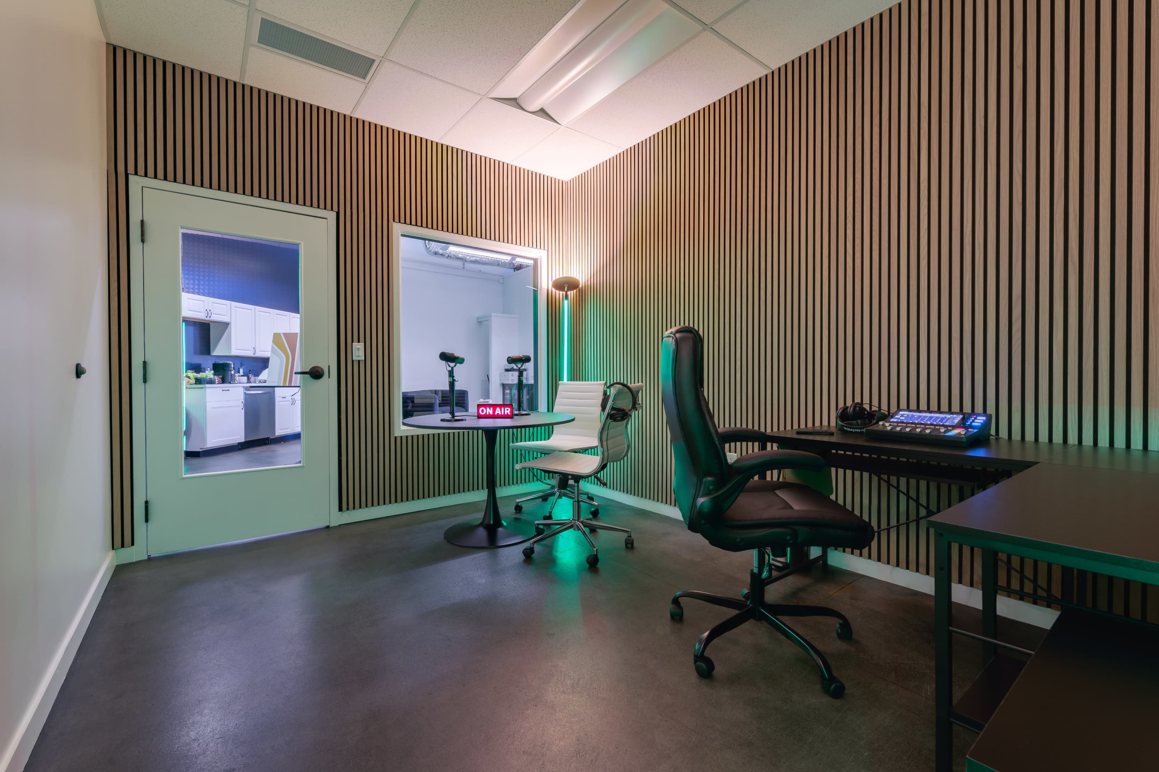 A modern recording studio with two chairs, a table, and audio equipment, along with a window viewing a separate space.