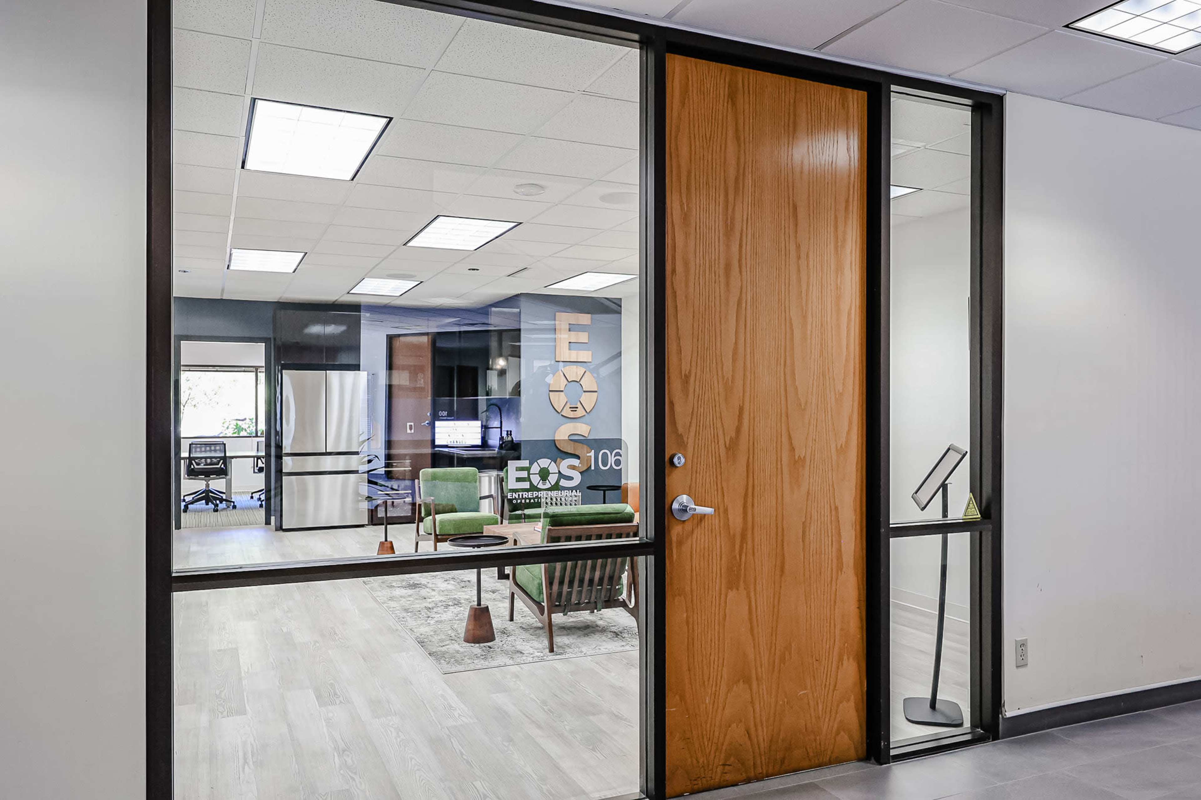 The image shows an office space with a glass door labeled "EOS 106," featuring a modern interior with seating and a kitchenette visible through the window.