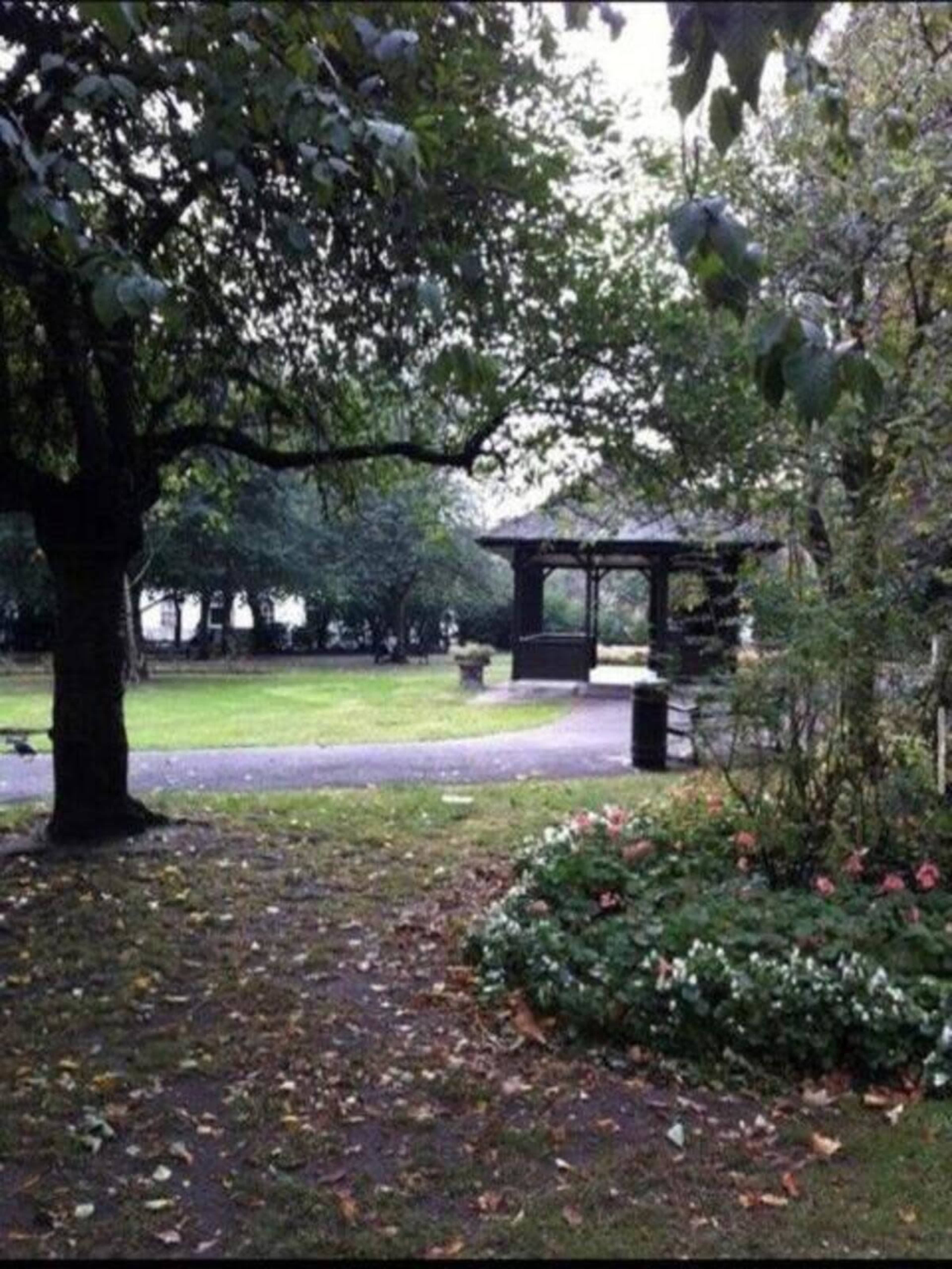 A park with lush green trees, a gazebo in the background, and a flower bed in the foreground.