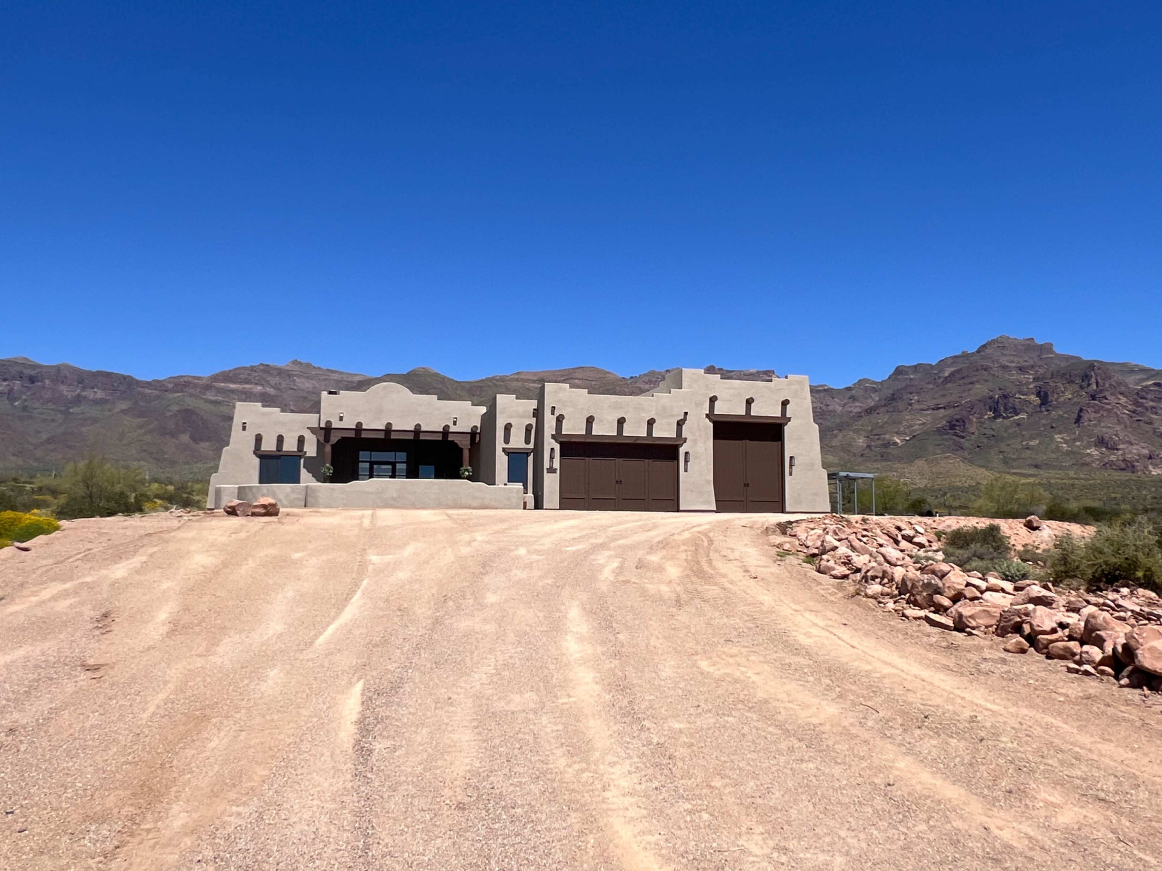 A large, adobe-style house with a flat roof and two garage doors sits at the end of a dirt driveway surrounded by mountains.