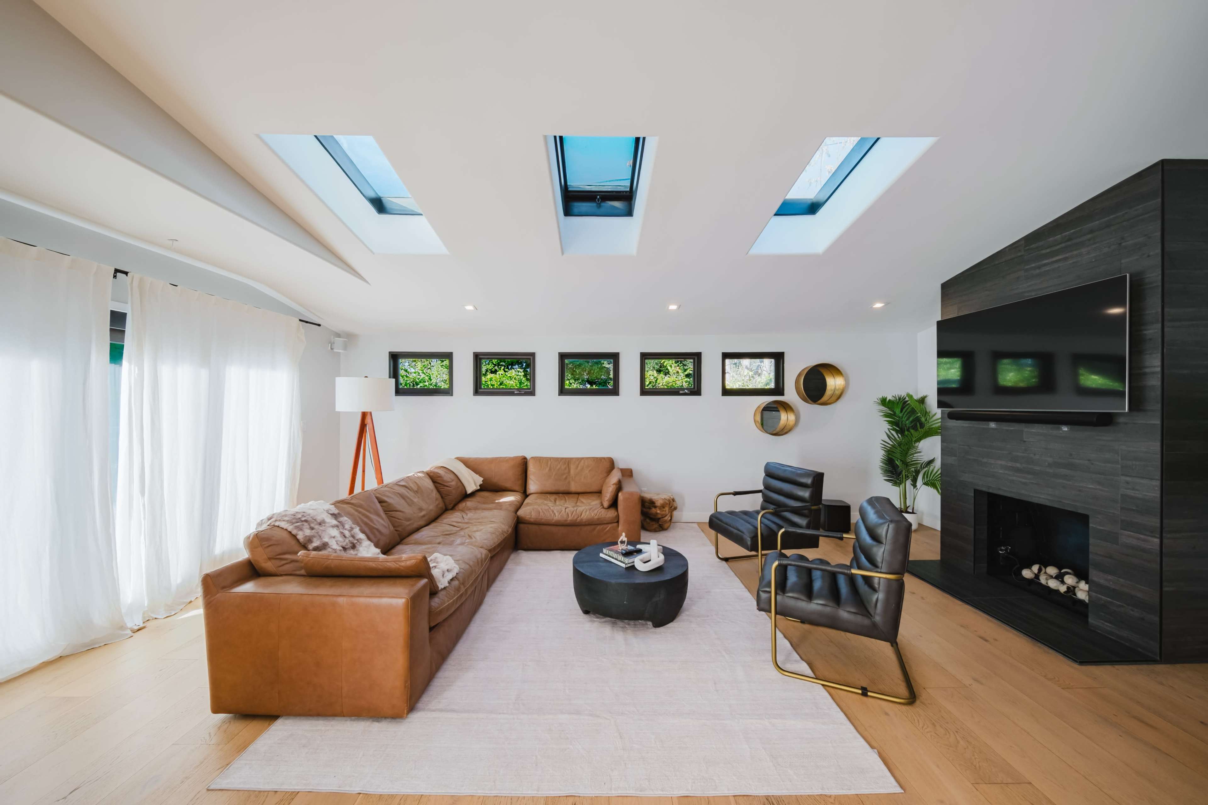 The living room features a large leather sectional sofa, black armchairs, a round coffee table, and a stone fireplace with a mounted television, illuminated by natural light from skylights.