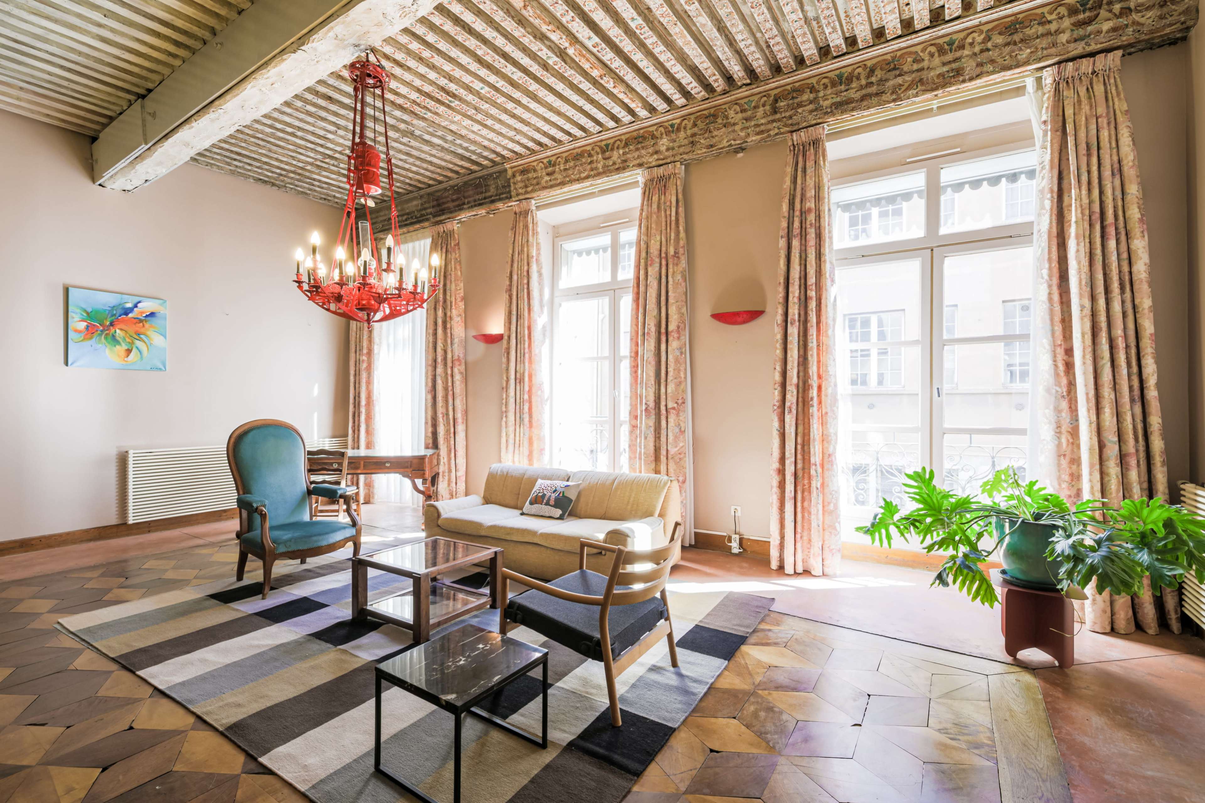 The image depicts a room with a chandelier, a sofa, and large windows letting in natural light, framed by patterned curtains and a decorative rug on the floor.