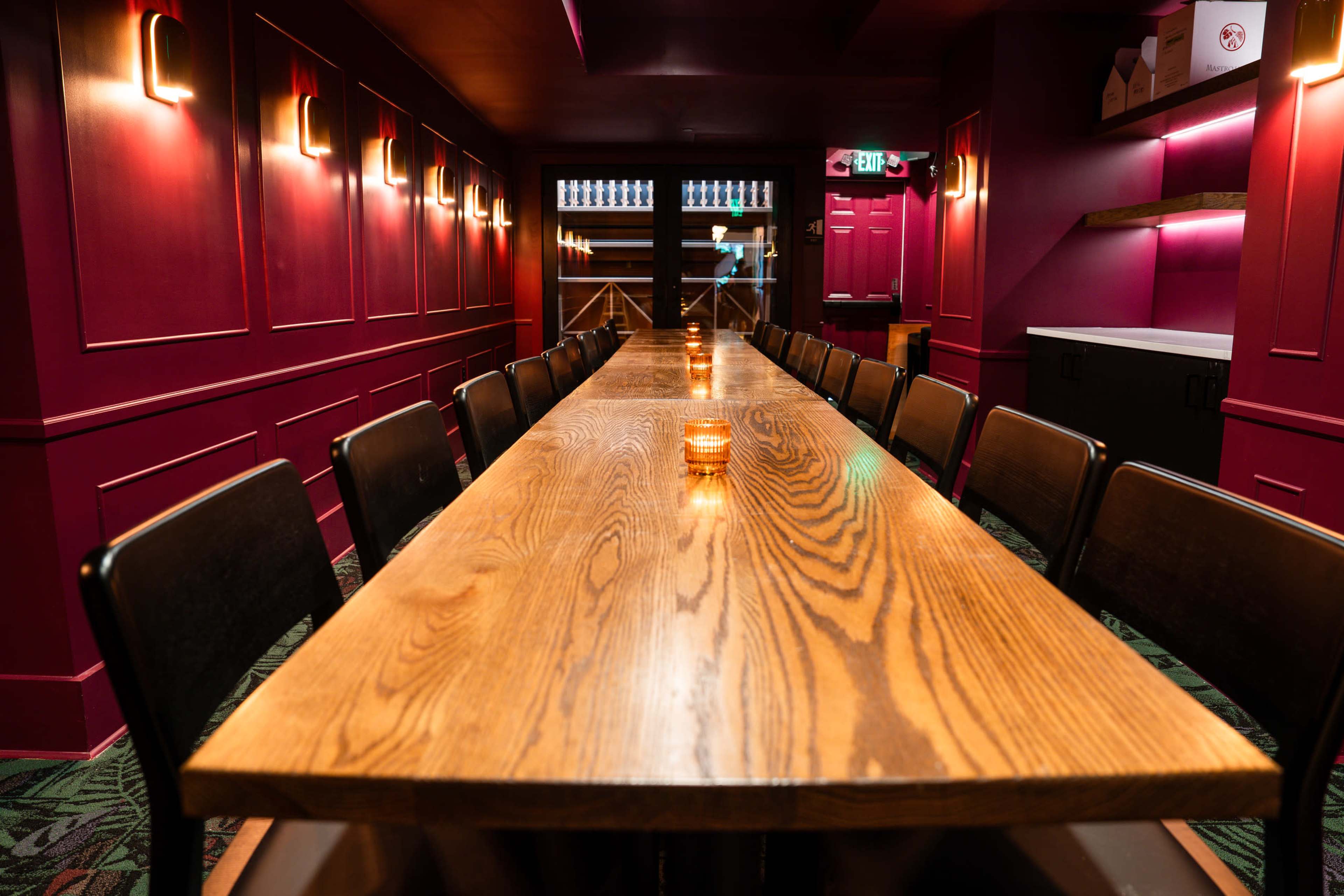A long wooden dining table with candles is set along a deep red wall in a dimly lit room.