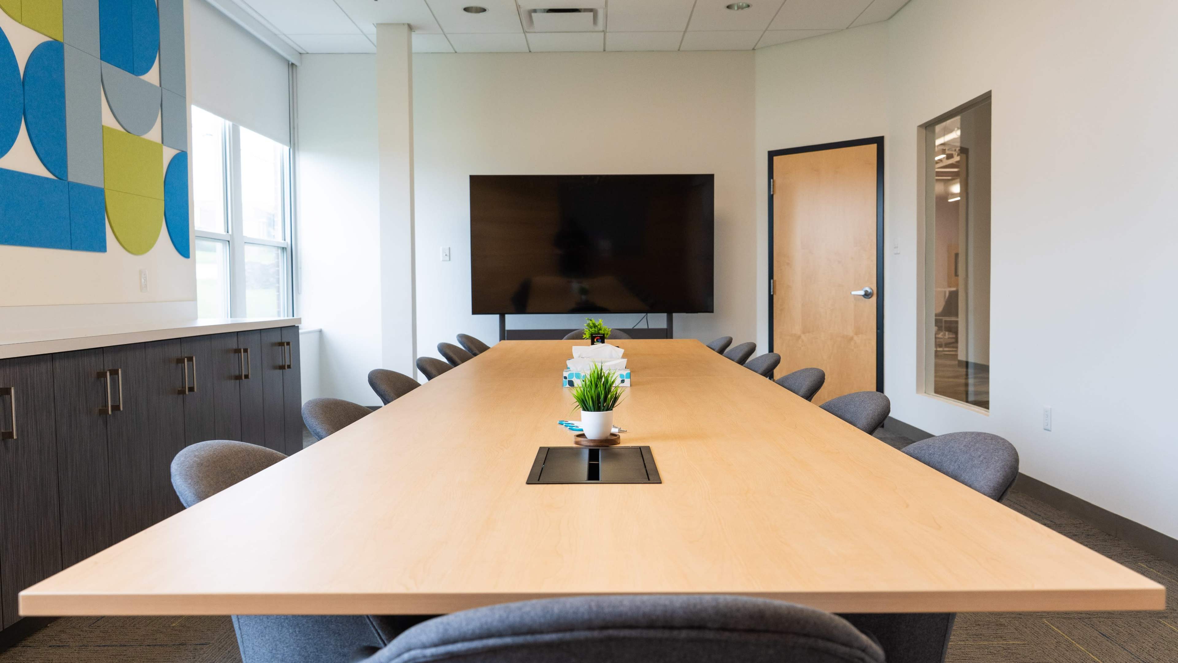A long wooden conference table with gray chairs surrounds a large meeting room featuring a wall-mounted television and a cabinet with colorful wall decorations.