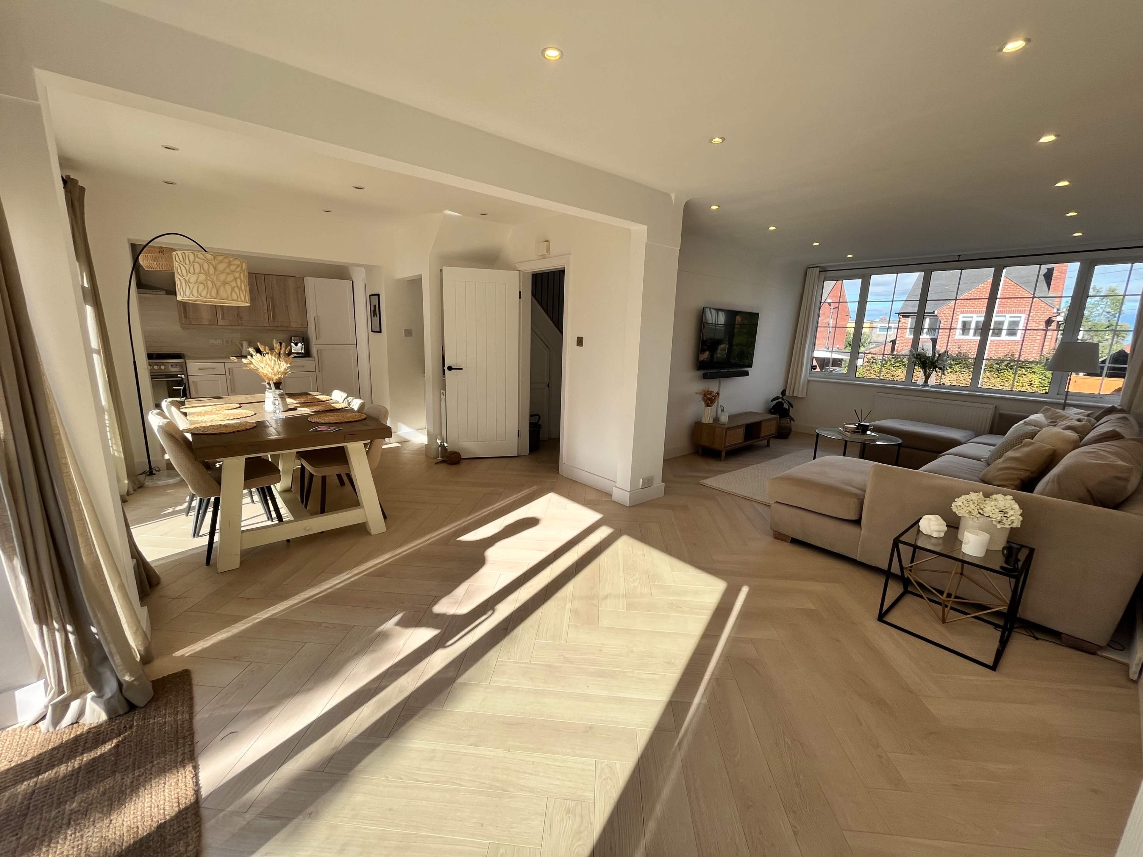 A modern open-plan living area with a dining table, a large sectional sofa, and large windows allowing natural light to fill the space.