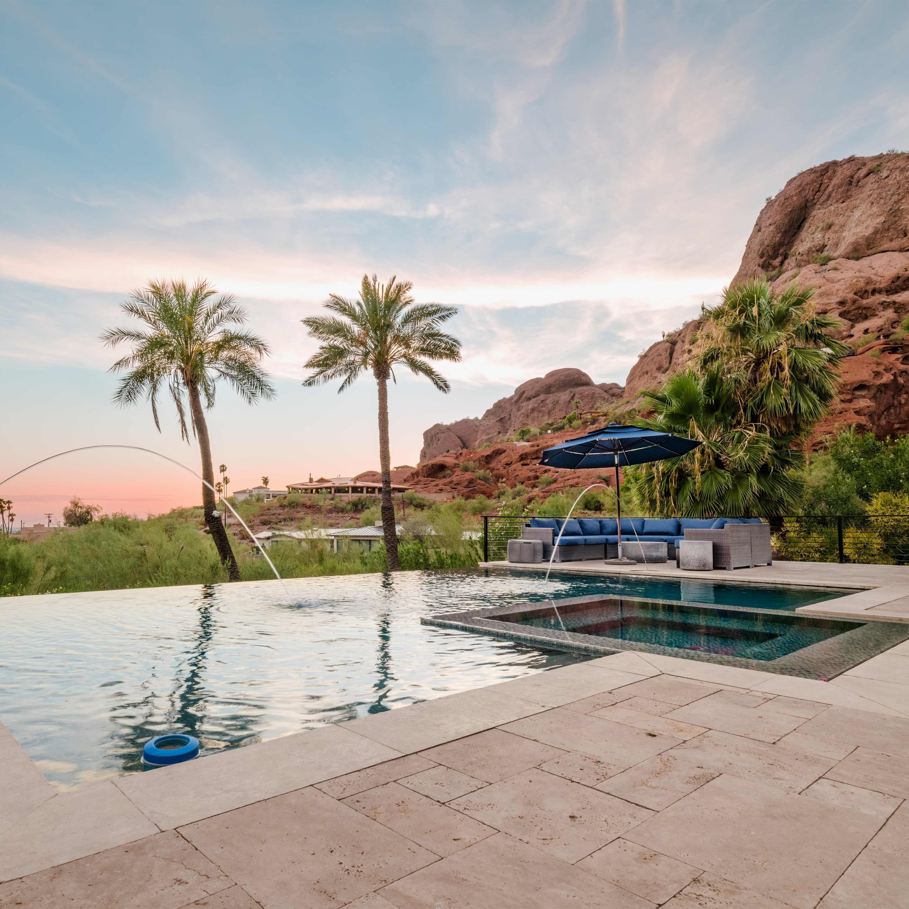 A swimming pool with an attached spa, surrounded by palm trees and set against a backdrop of rocky hills under a colorful sunset sky.