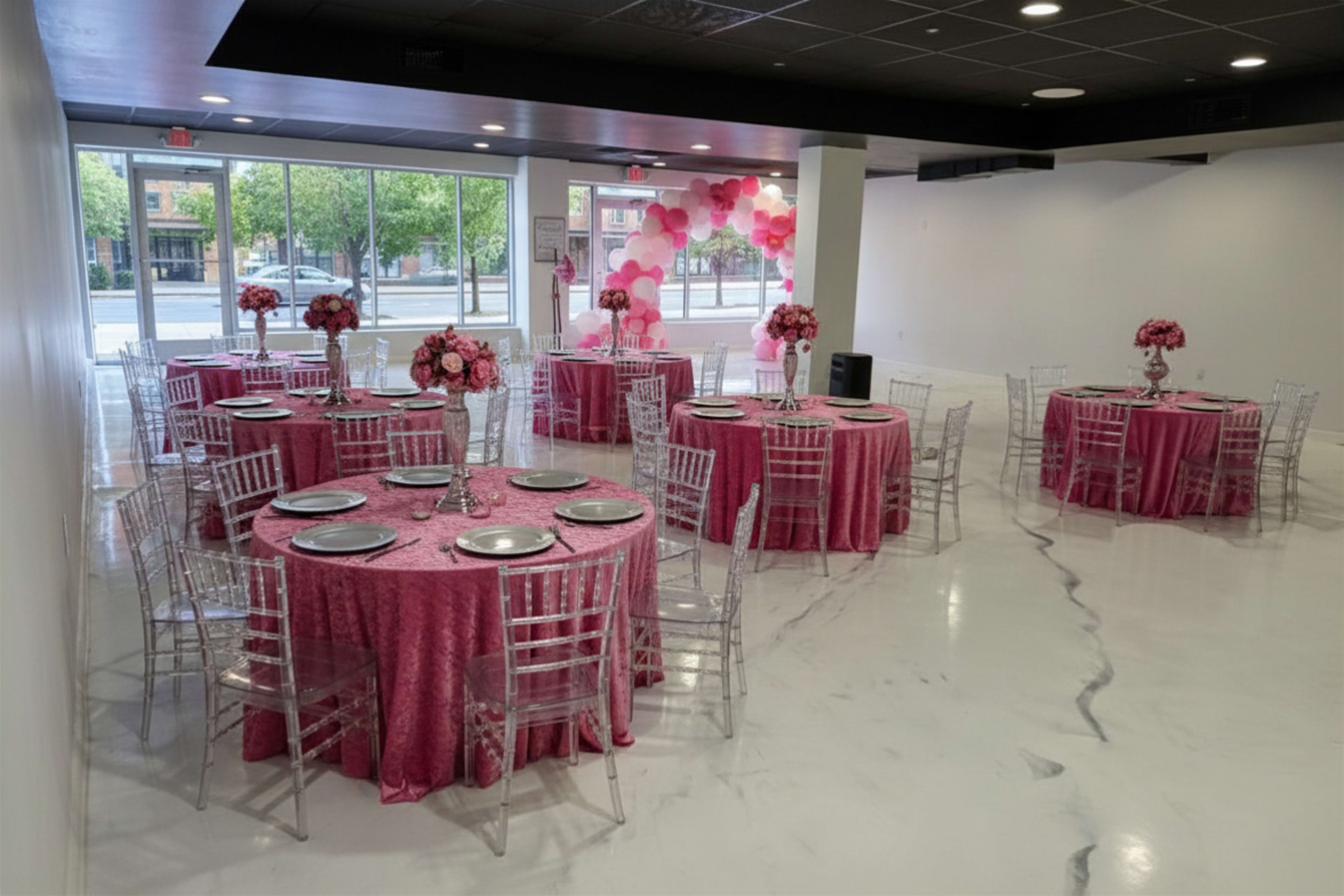 The image shows a decorated event space with several tables covered in pink tablecloths and adorned with floral centerpieces.