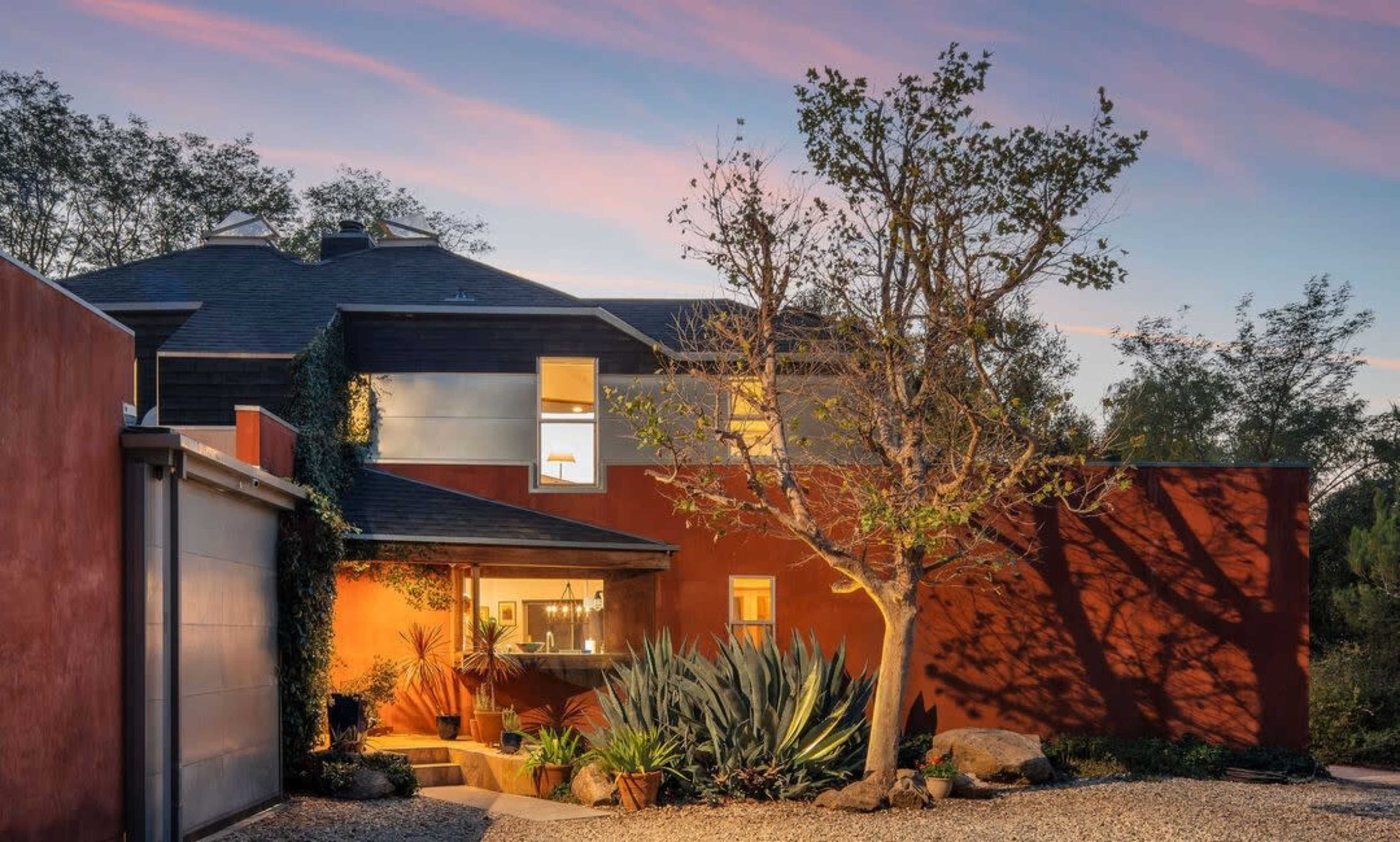 A modern house with a warm-lit entrance, surrounded by landscaping featuring plants and a prominent tree against a twilight sky.