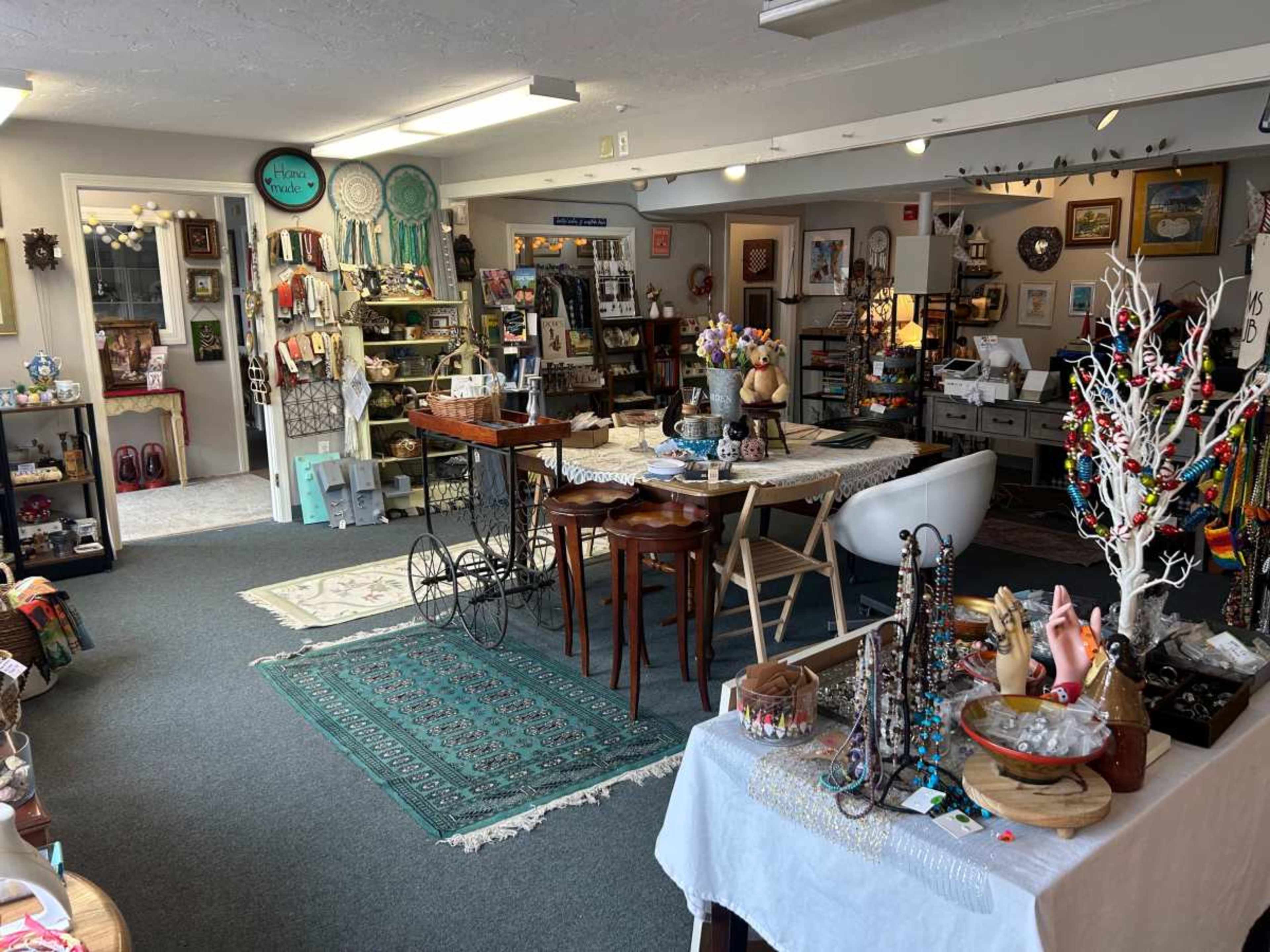 The image shows a cozy shop interior filled with various crafts, decorations, and handmade items, featuring tables, displays, and colorful merchandise.