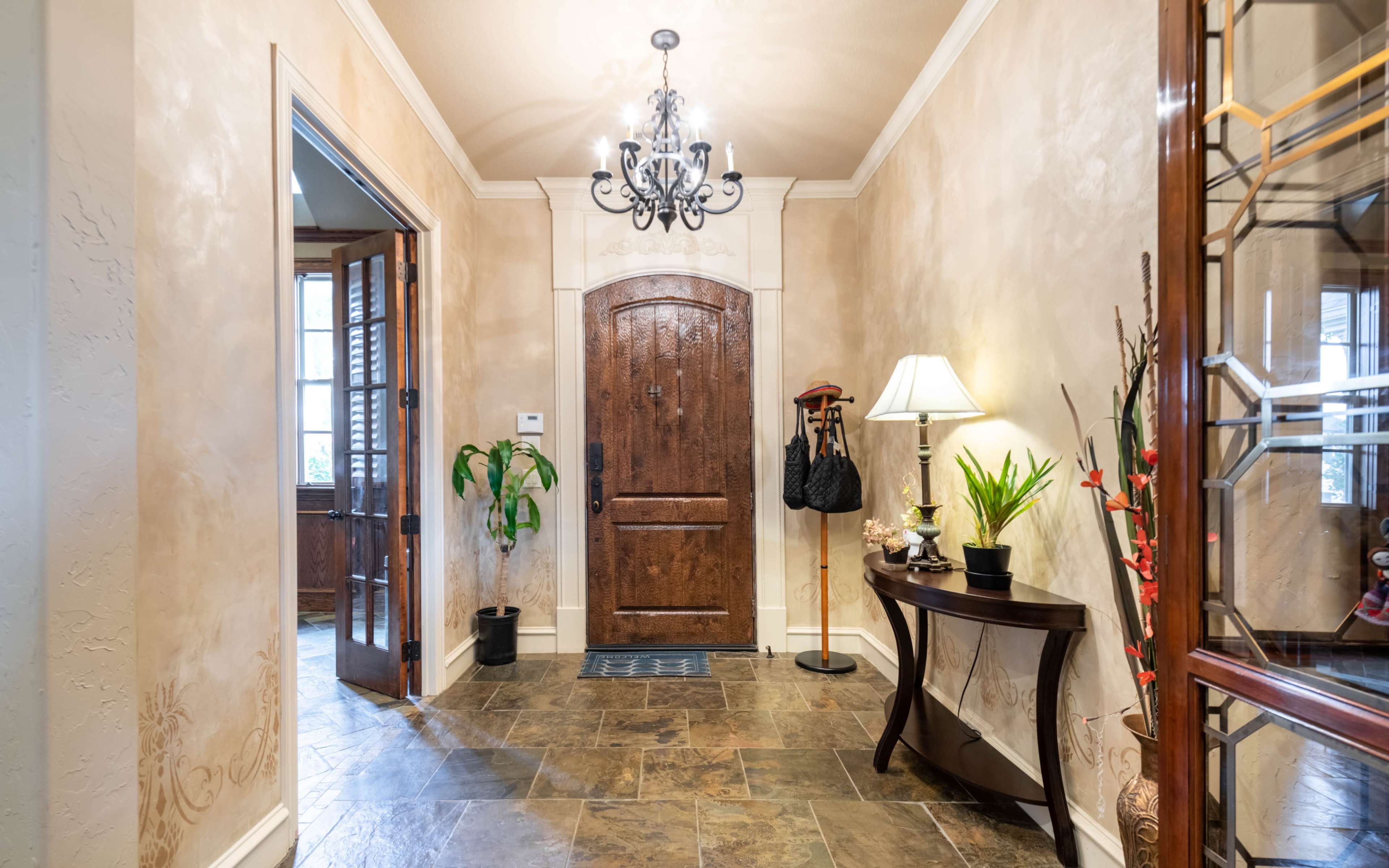 A well-decorated entryway featuring a wooden front door, a chandelier, a console table with decorative plants, and a lamp beside it.
