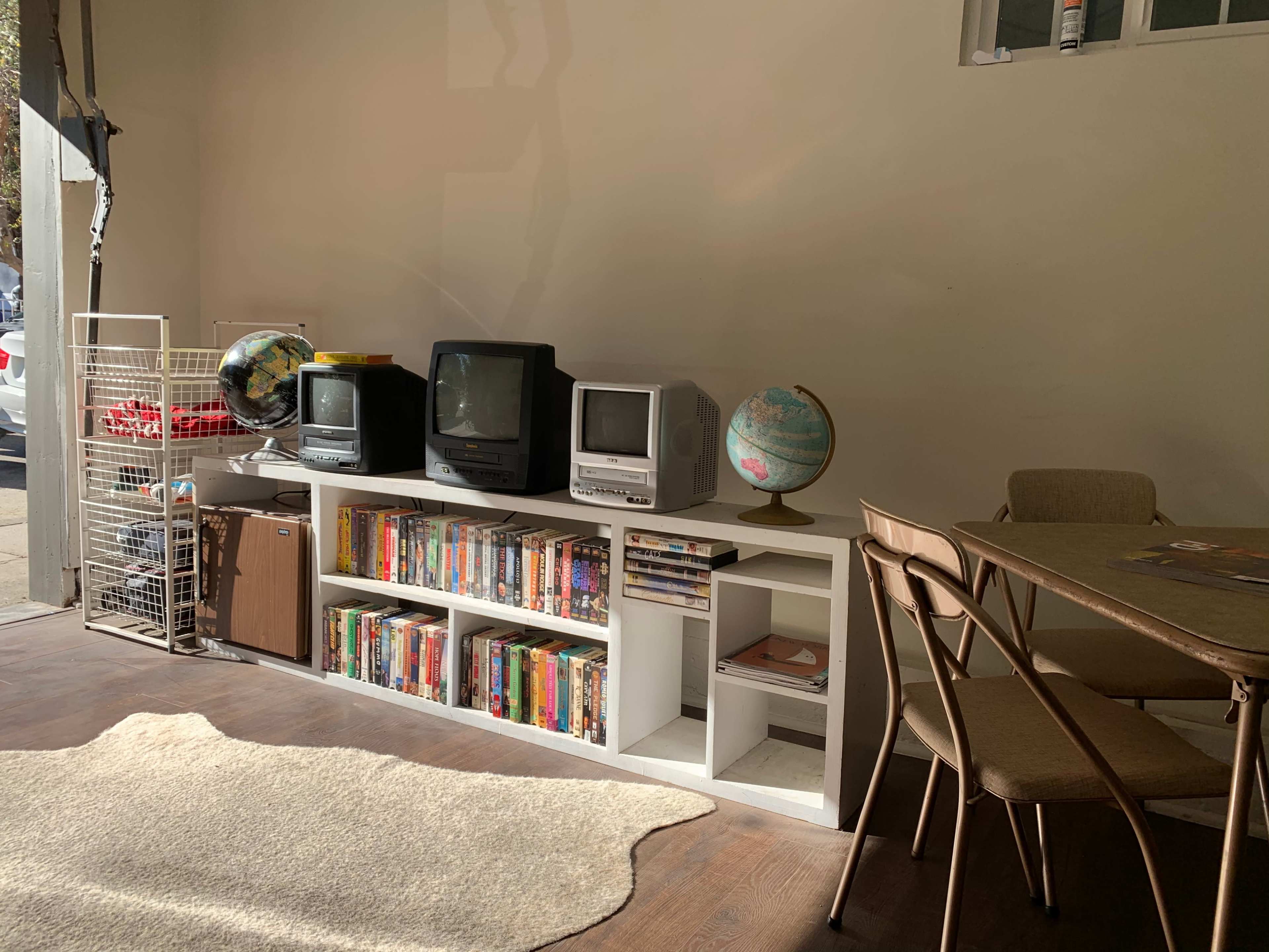 A room with two old televisions, a globe on a table, a bookshelf filled with DVDs, and a dining area with chairs.