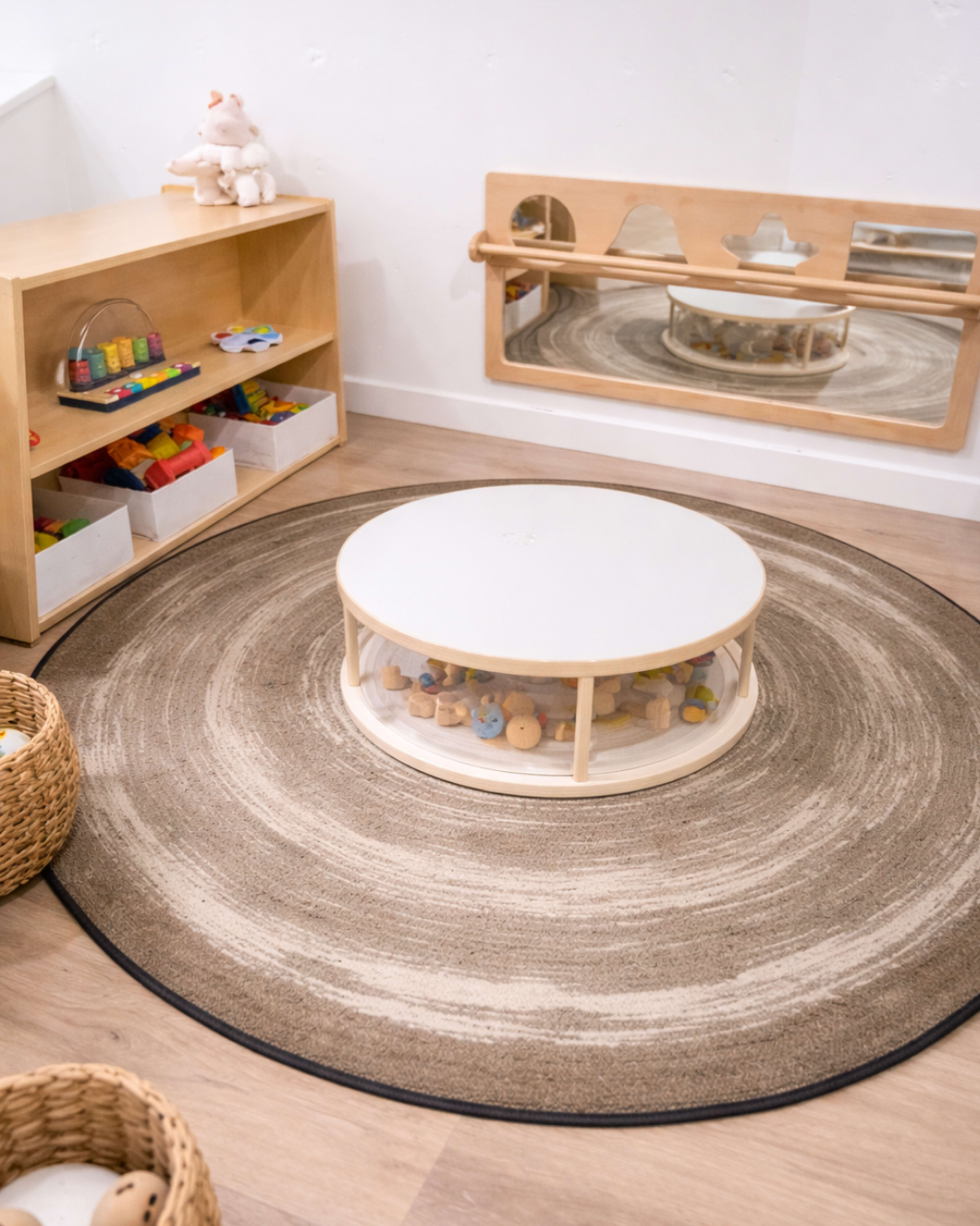 The image shows a children's play area featuring a circular rug, a low table with a transparent container of toys, wooden storage shelves filled with colorful toys, and a wall-mounted mirror.