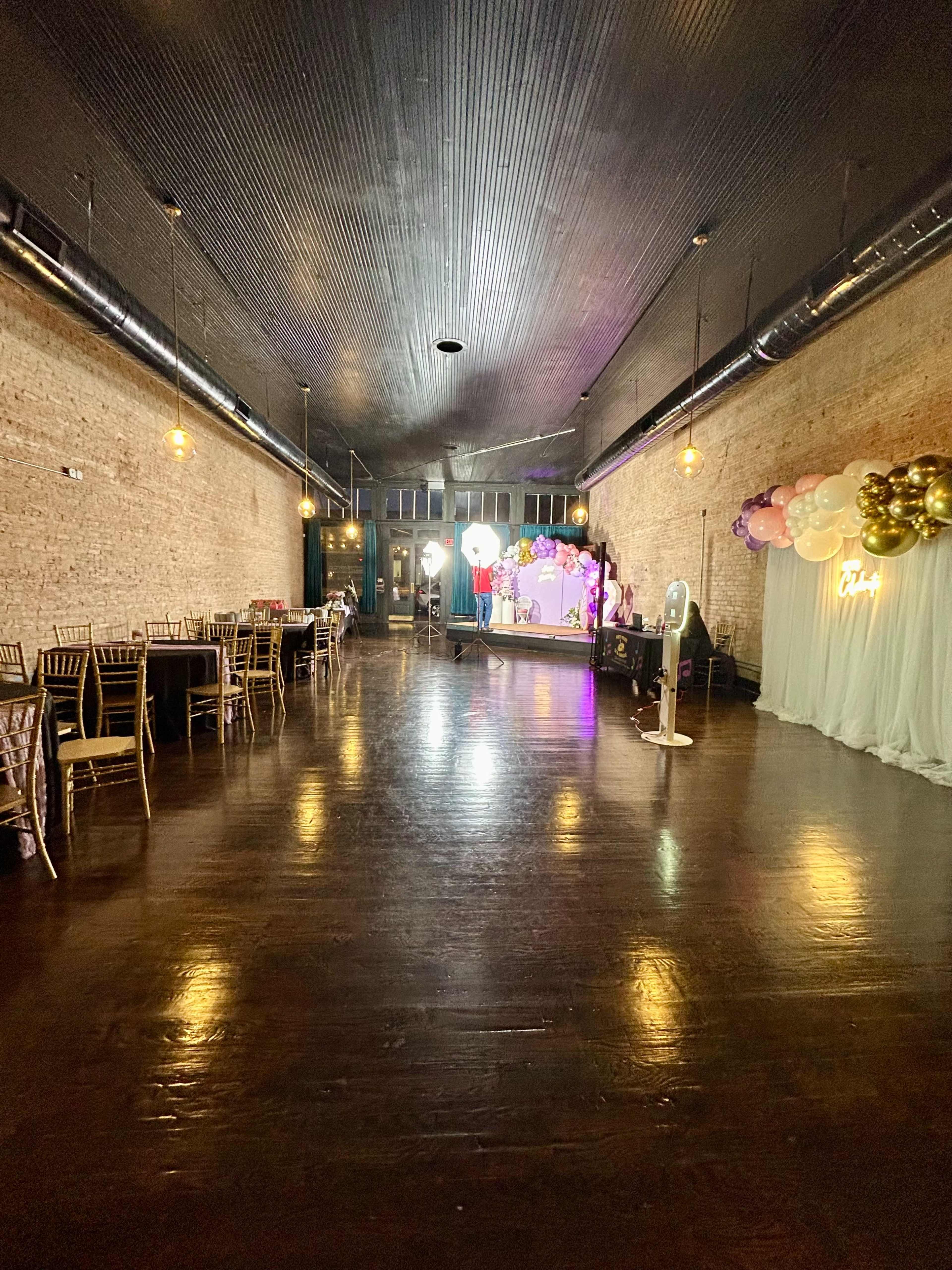 A spacious event hall features wooden floors, exposed brick walls, and a staged area decorated with balloons and a backdrop.