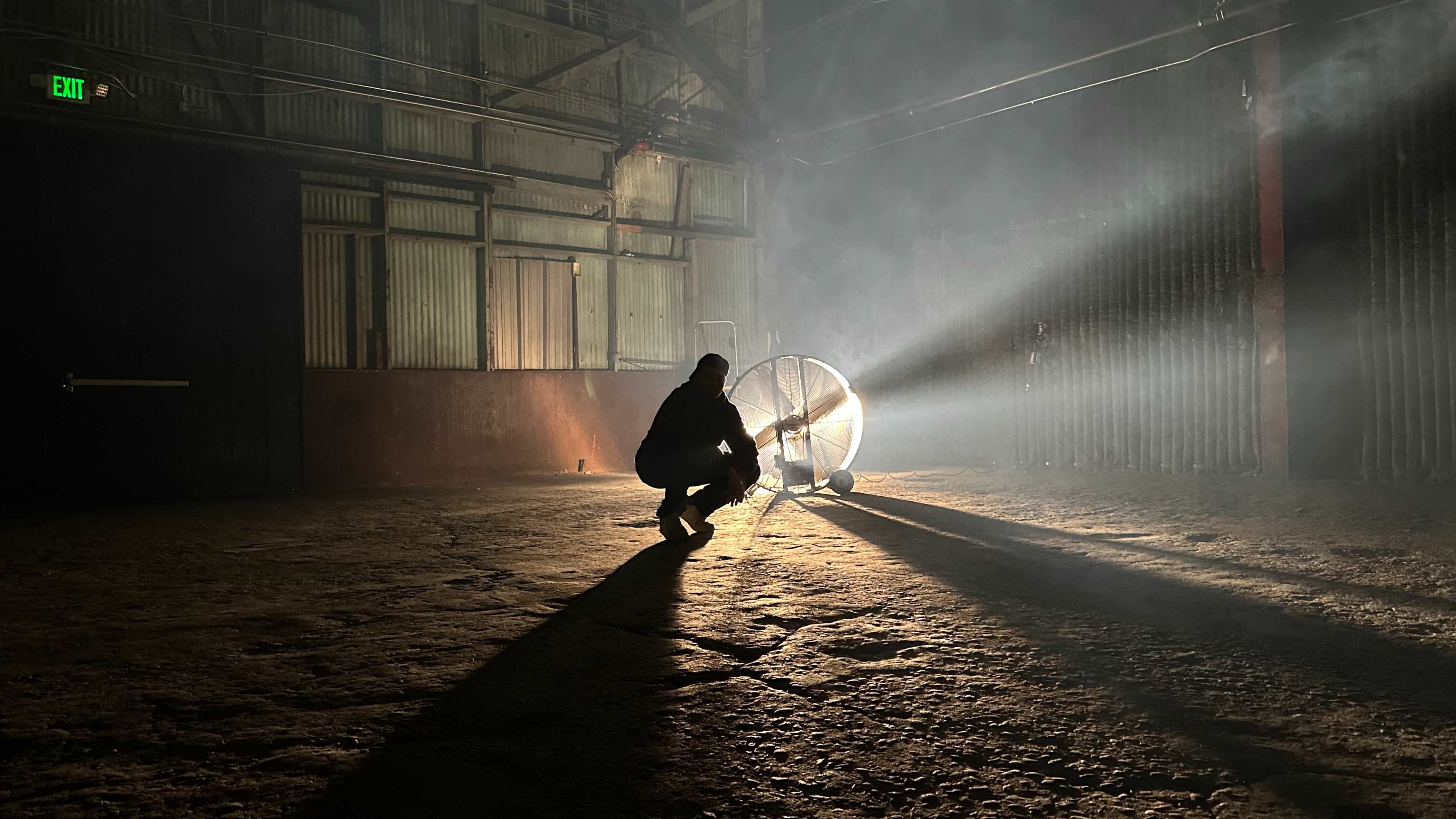 A person in a dark, industrial space crouches near a large fan, casting long shadows and beams of light through the mist.