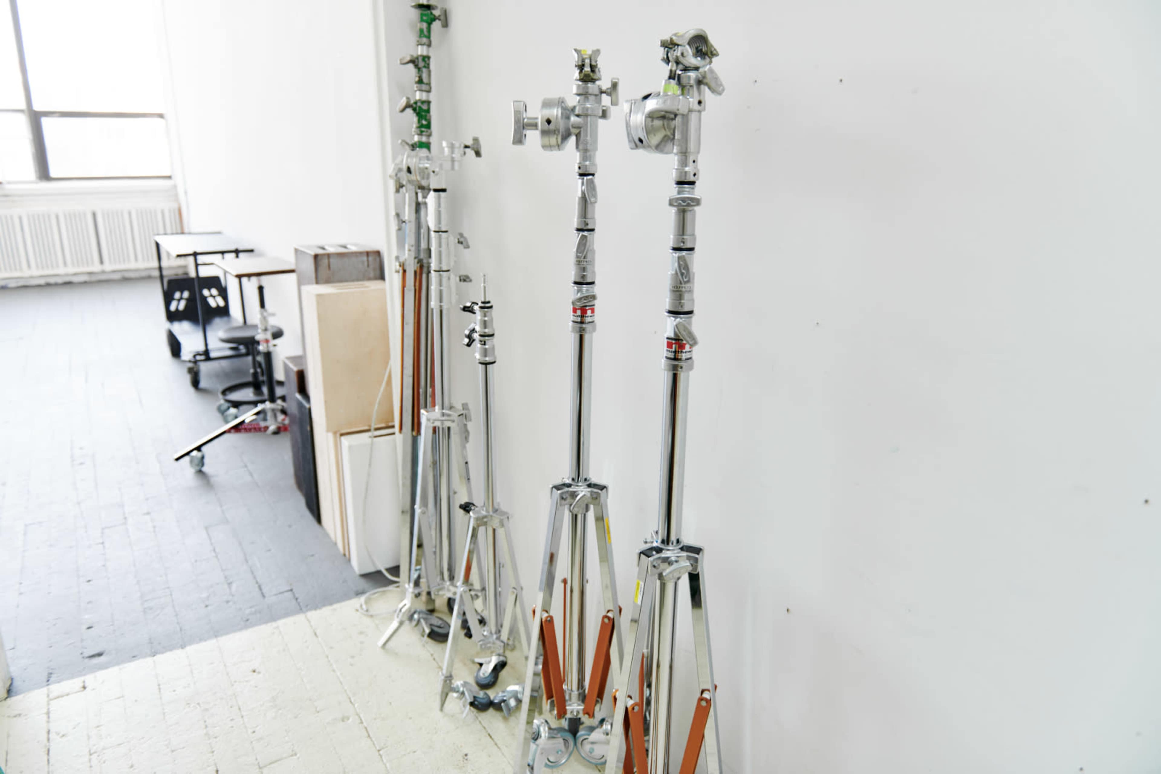 Several aluminum light stands with adjustable heights are arranged along a white wall in a studio space.