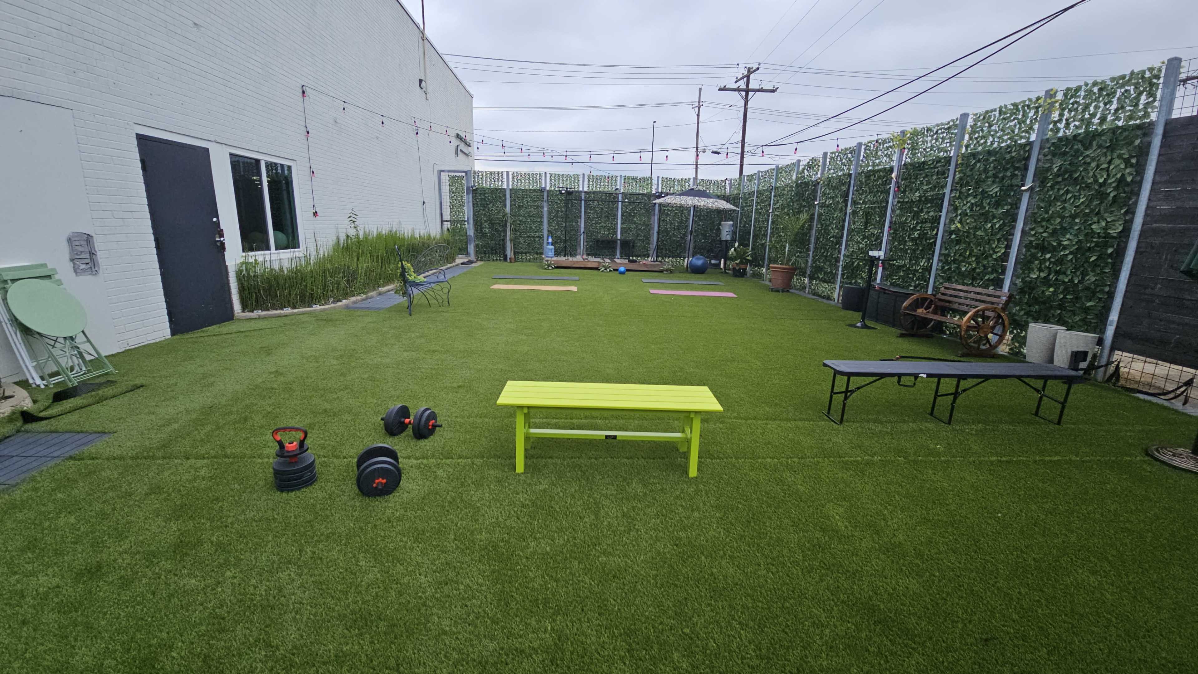 The image shows an outdoor fitness area with artificial grass, featuring exercise equipment such as weights and benches, surrounded by greenery and buildings.