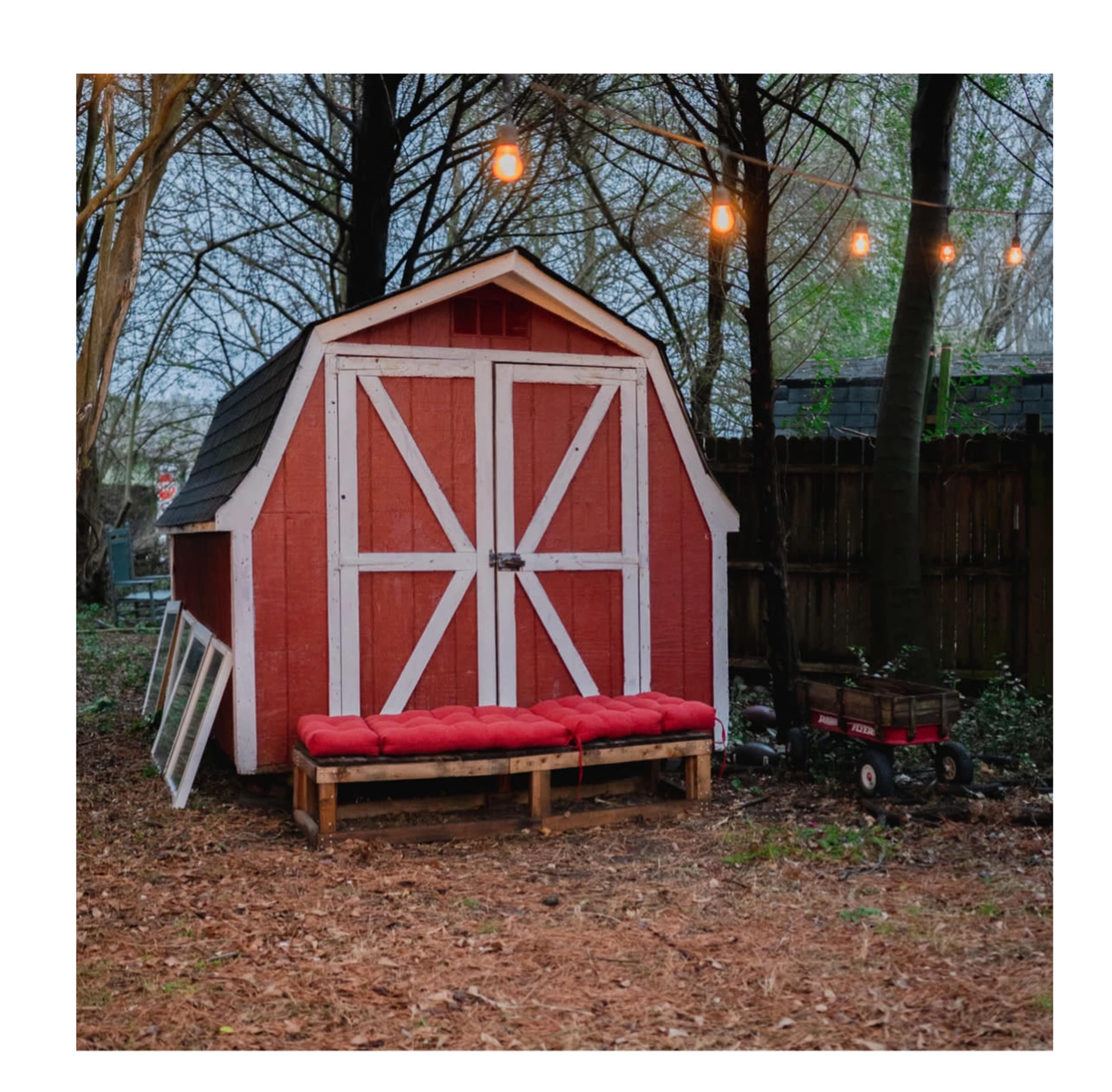 A red barn-style shed with white trim and a wooden bench sits in a wooded area illuminated by string lights.