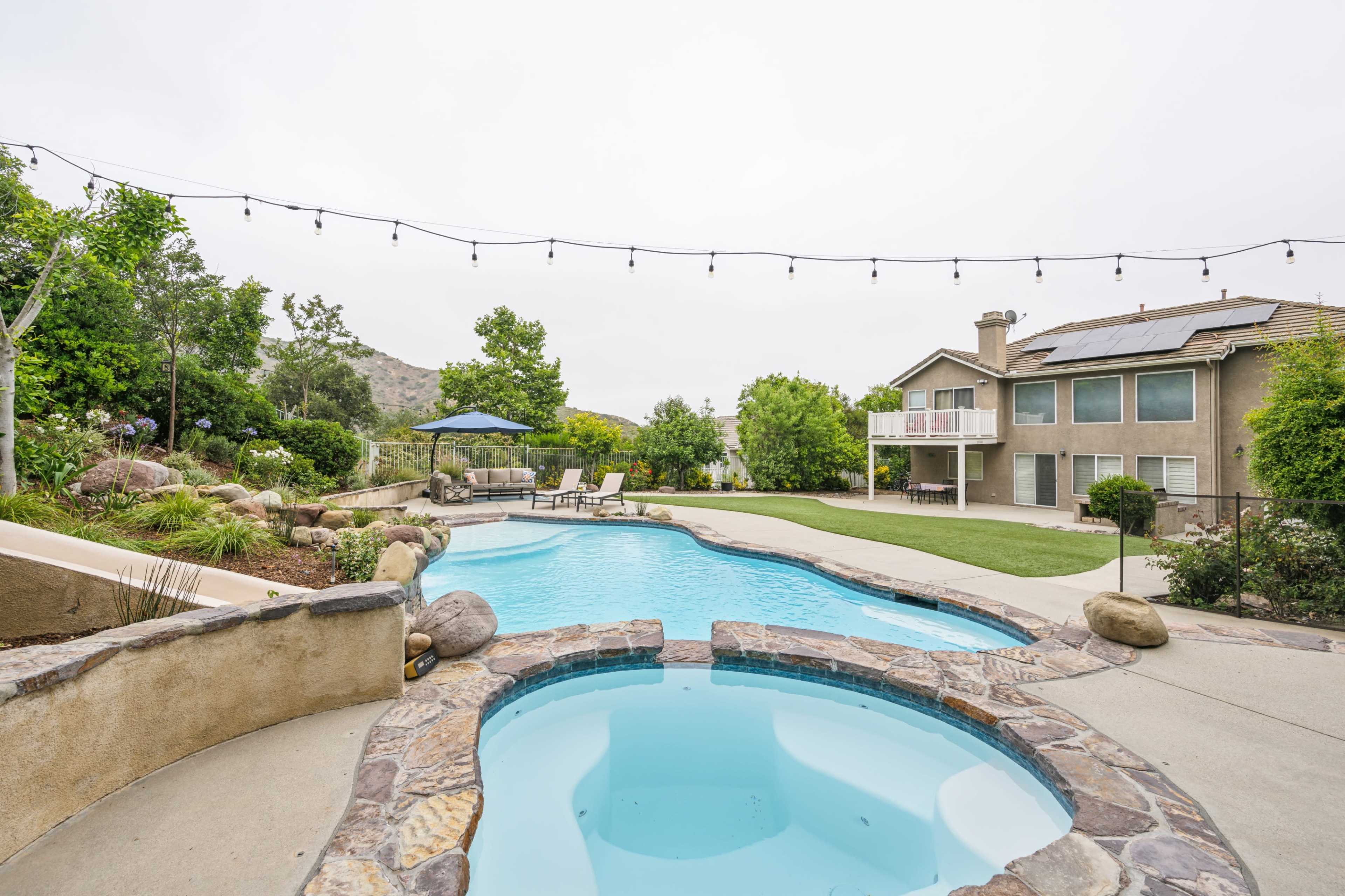 A backyard with a swimming pool and spa, surrounded by landscaping and a view of a hillside in the background.