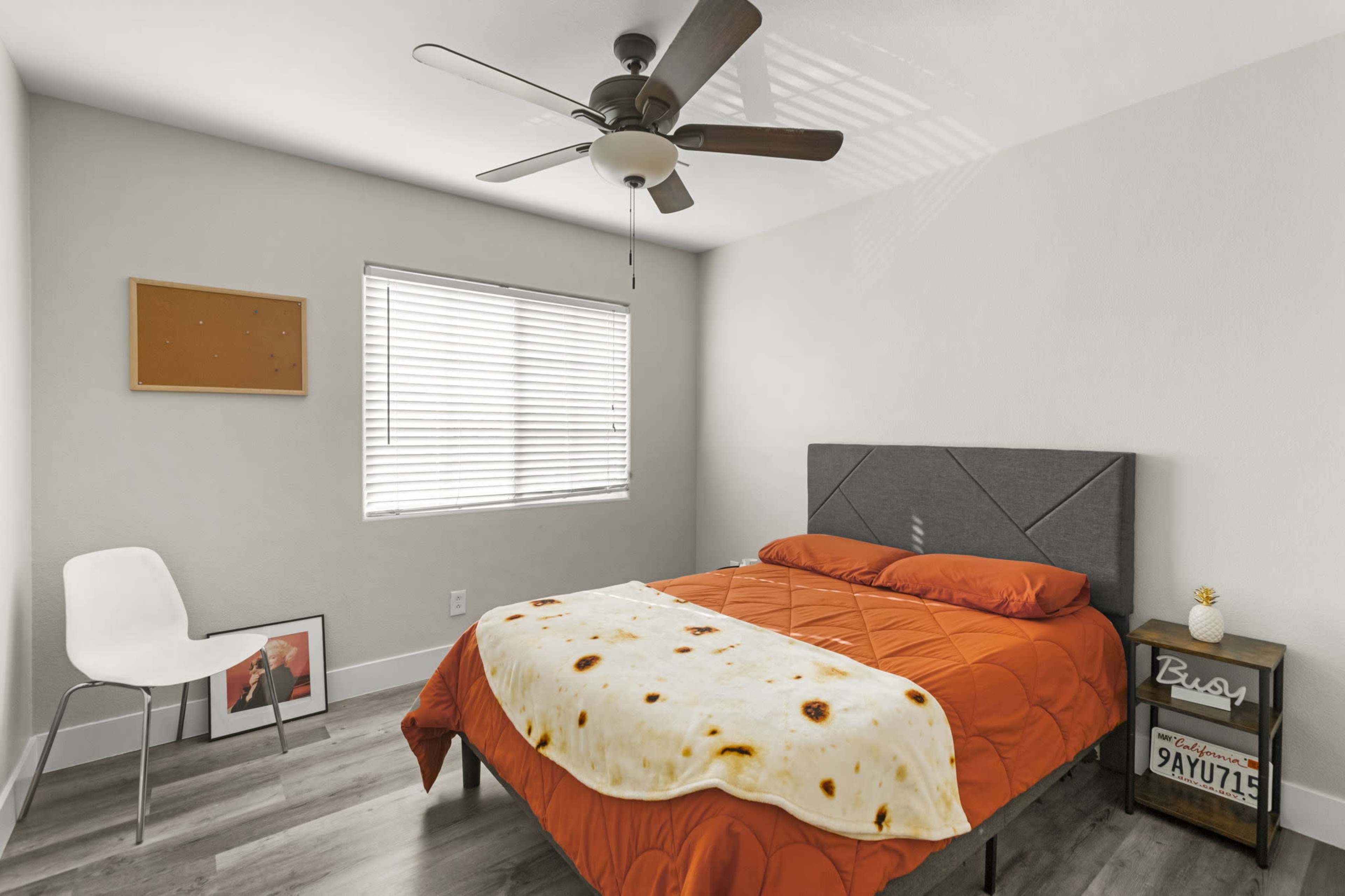 The image shows a small bedroom with an orange bedspread, a ceiling fan, a window with blinds, a white chair, and a nightstand with a framed picture.