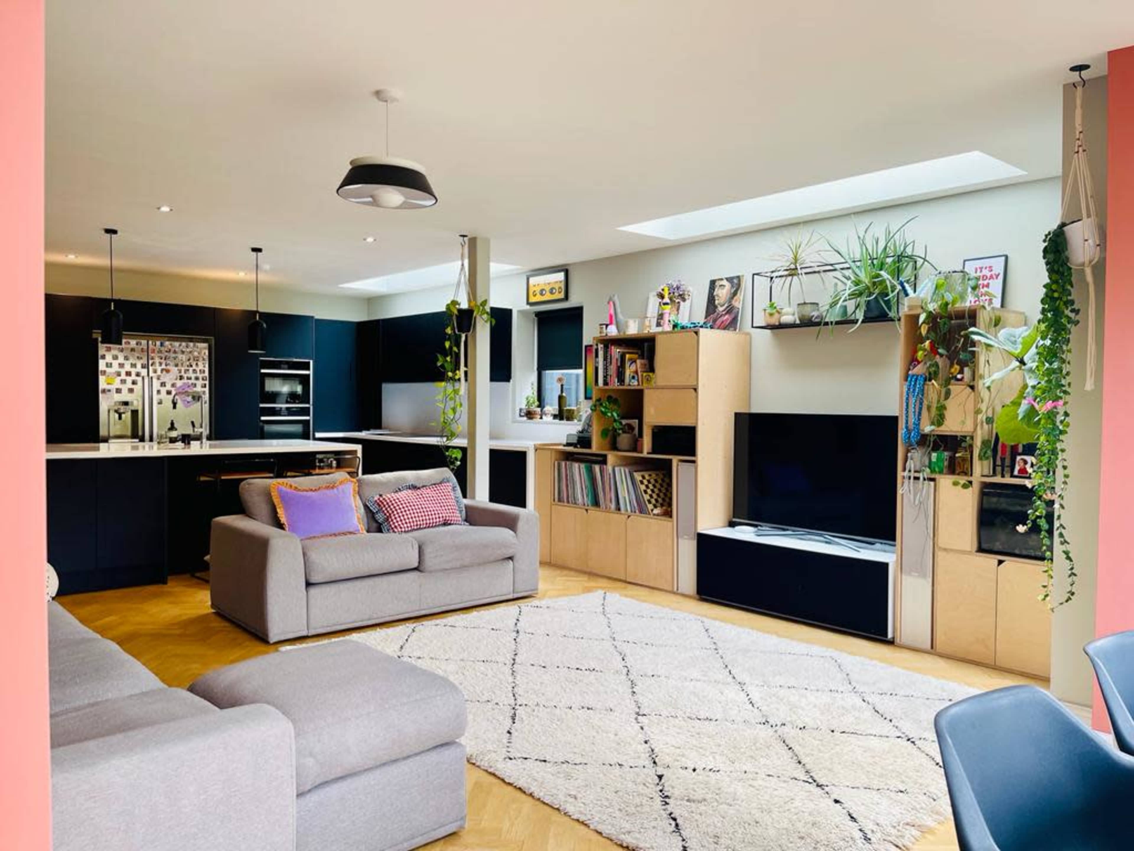 The image shows a modern living room with a kitchen, featuring a sofa, a television unit, shelves filled with books and plants, and a large area rug.