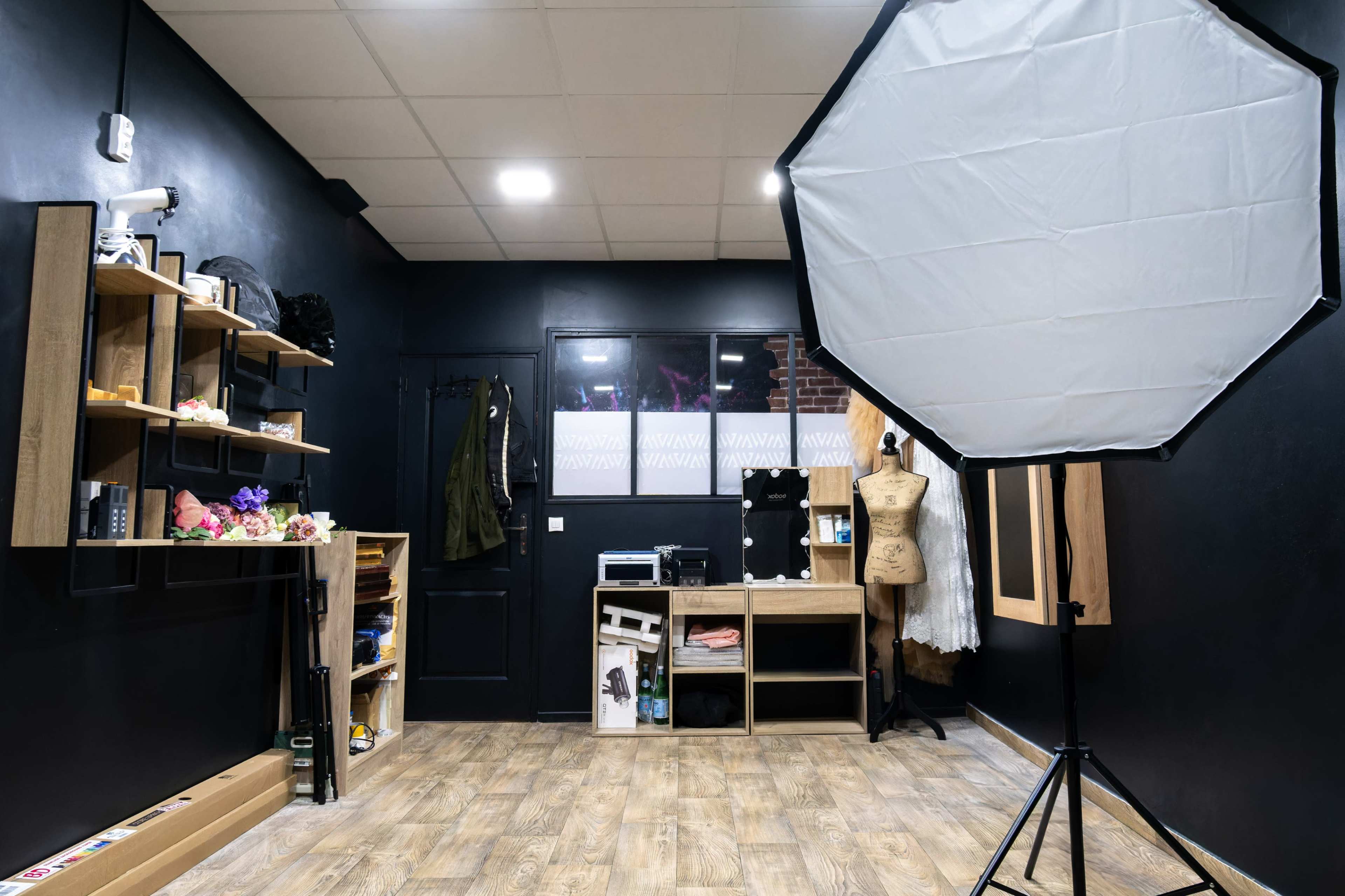 The image shows a photography studio with black walls, wooden shelving filled with various items, and a large softbox lighting setup in the foreground.