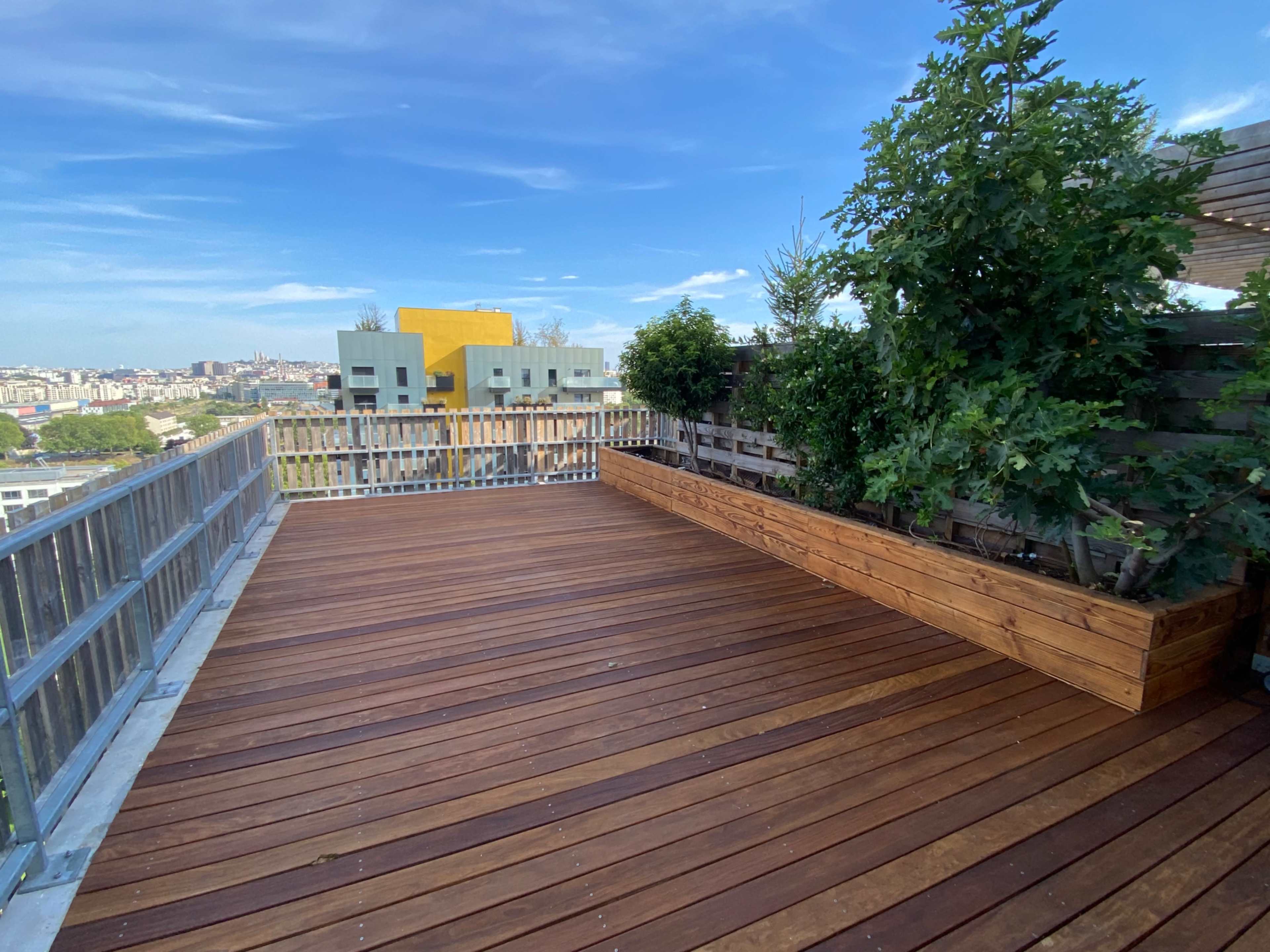 A spacious wooden terrace with planters and a panoramic city view under a clear blue sky.