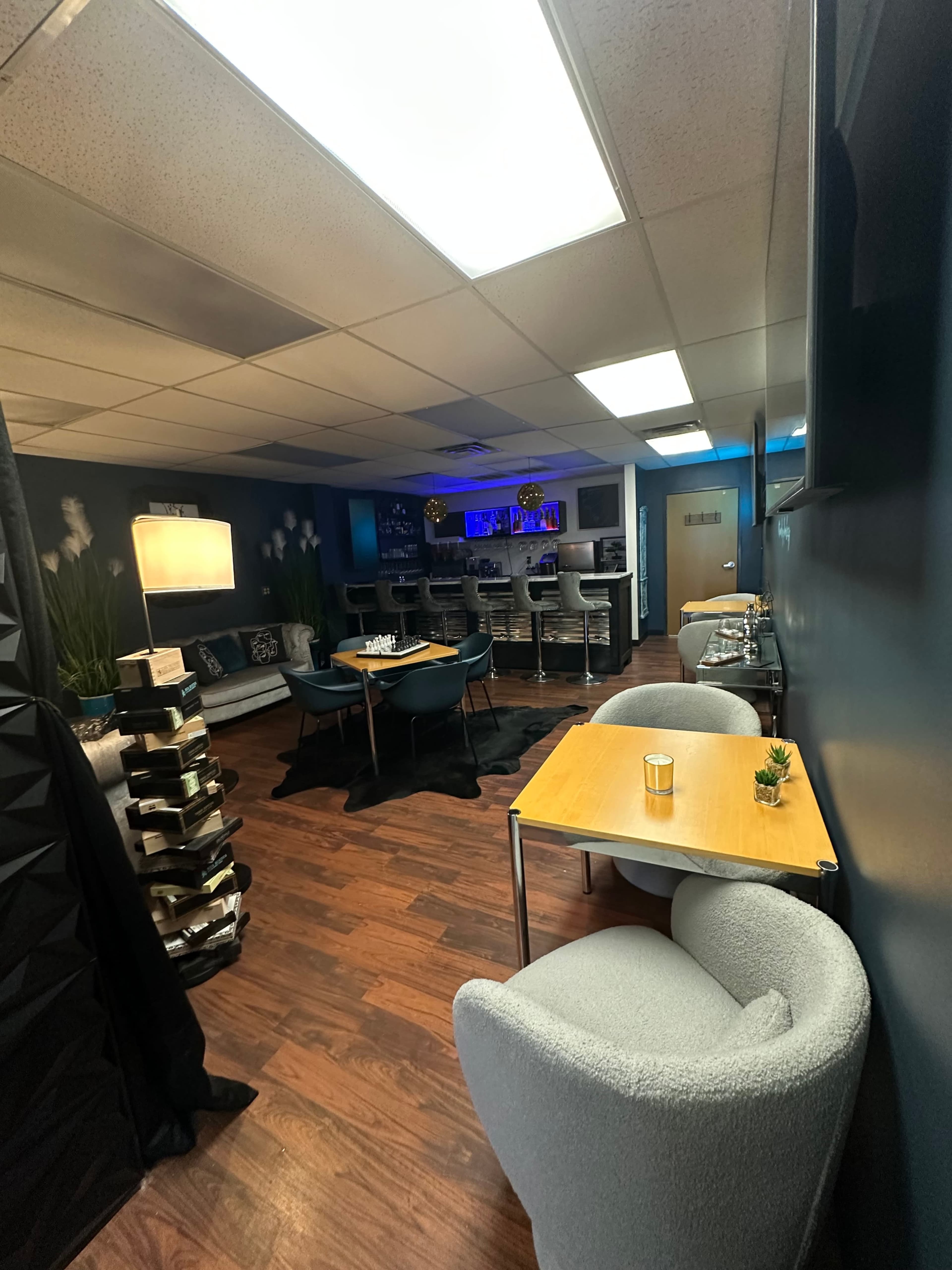 The image shows a modern lounge area with seating, tables, and a bar in a dimly lit space.