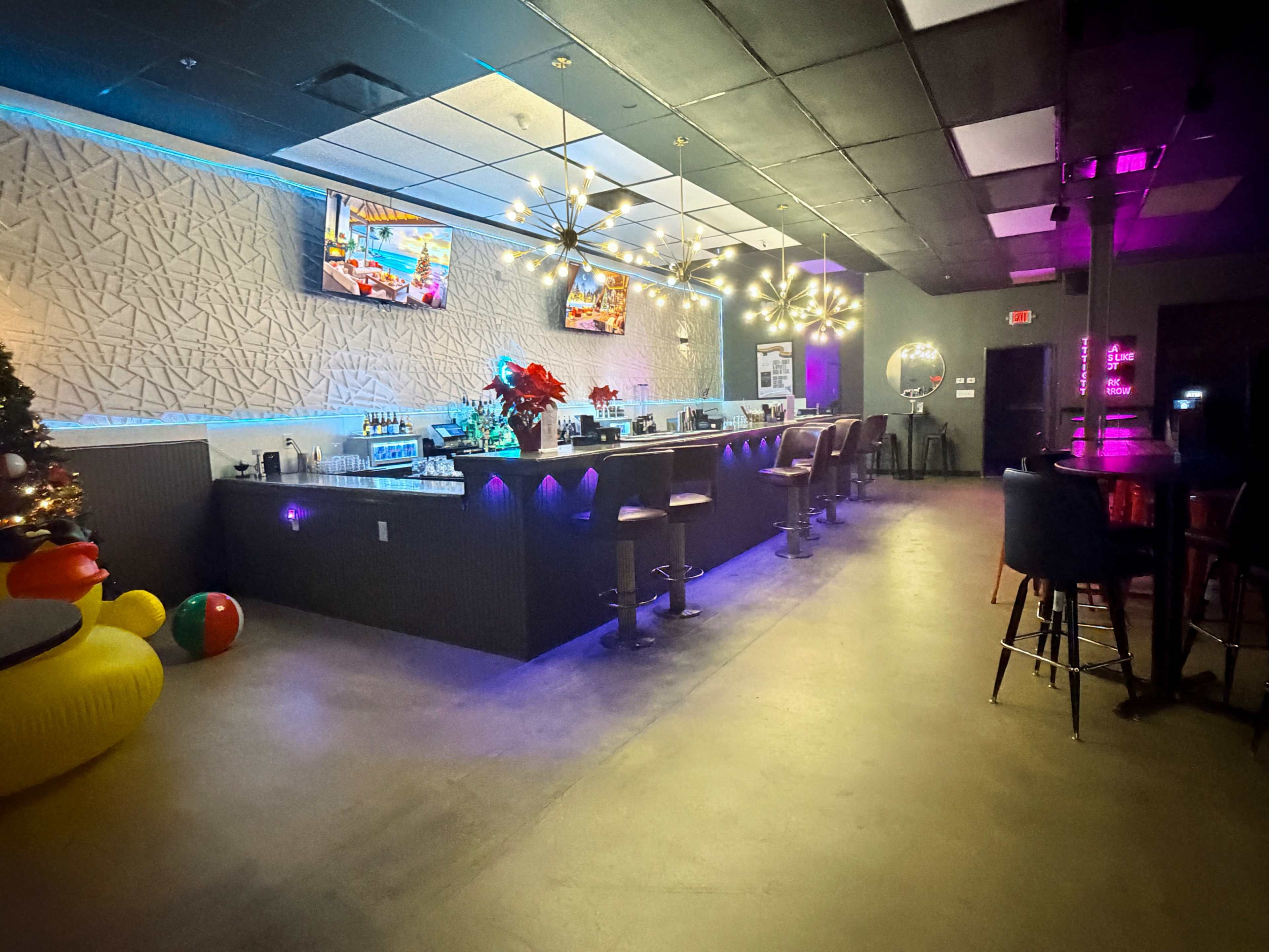The image shows a modern bar area with a long counter, colorful lighting, and several high stools, along with two televisions mounted on the wall.