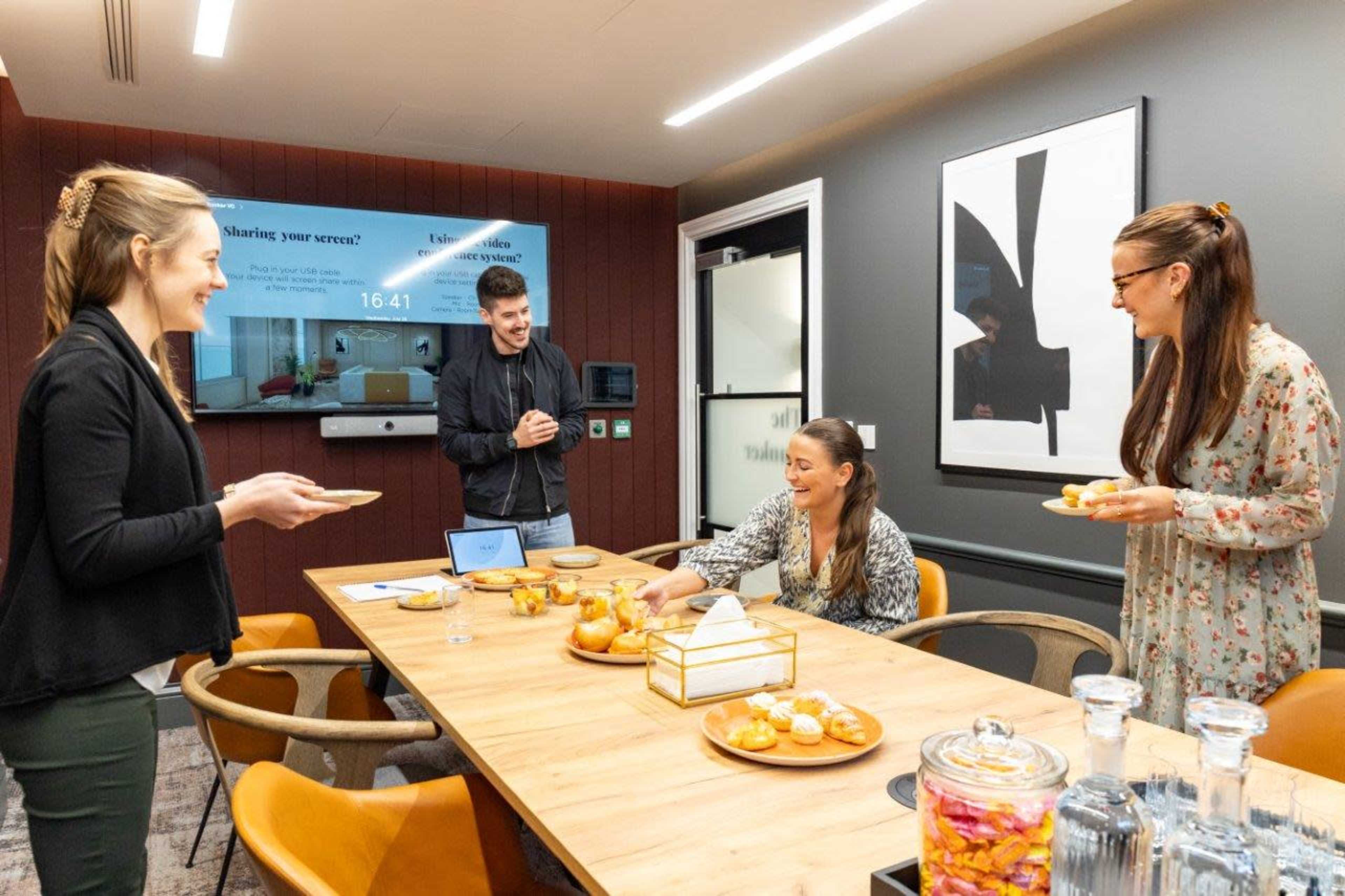 Four people are gathered around a conference table with snacks, engaging in conversation in a modern meeting room.