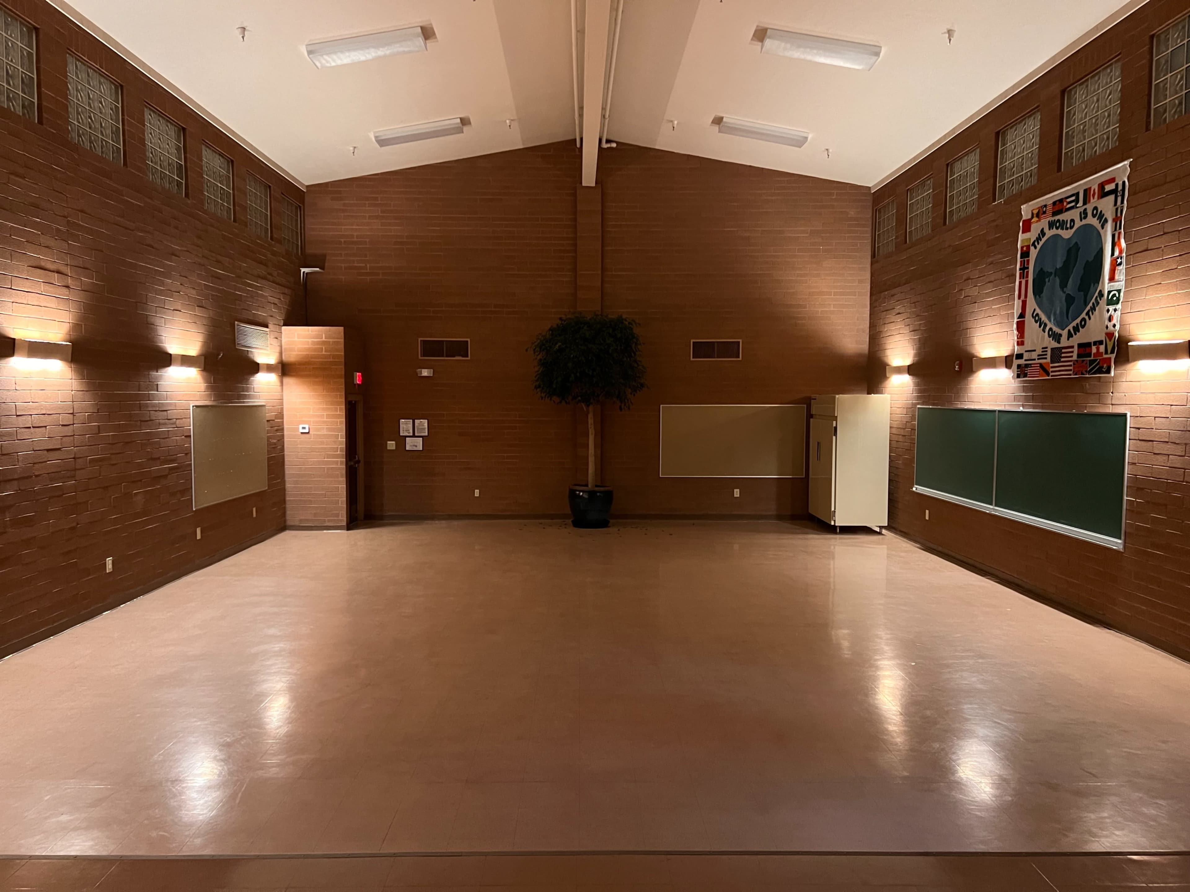 The image shows a spacious, empty room with a polished floor and brown brick walls, featuring a potted plant and two large whiteboards.