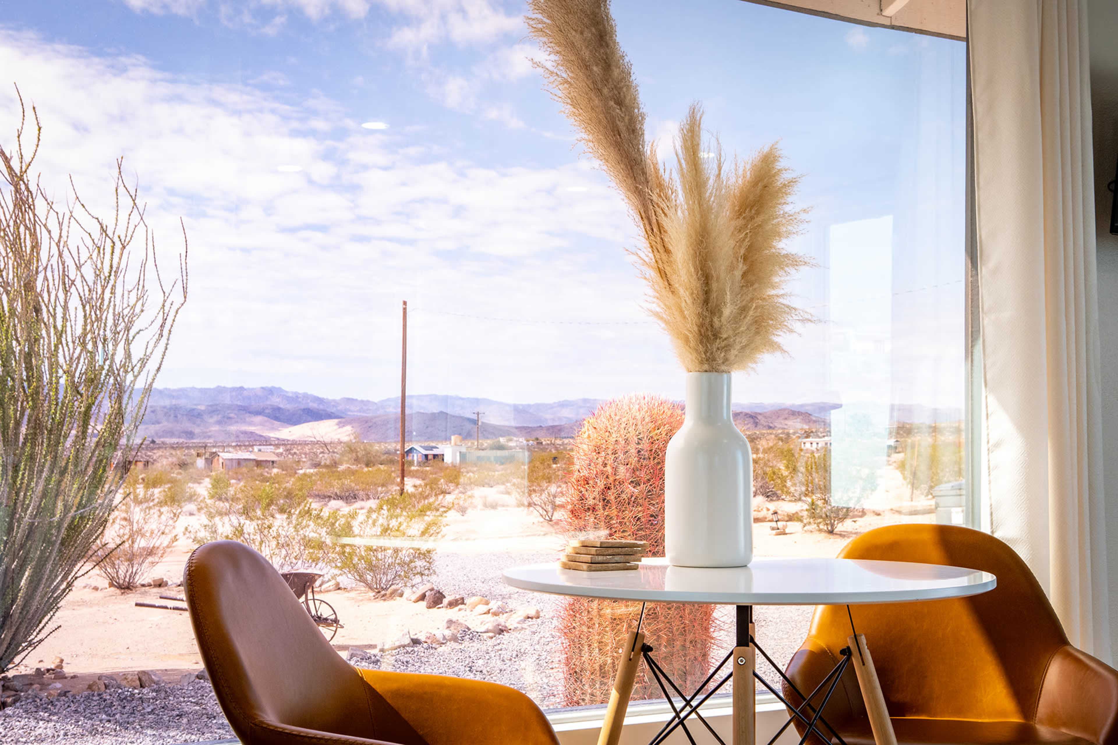 A white vase with pampas grass sits on a round table in front of large windows overlooking a desert landscape.