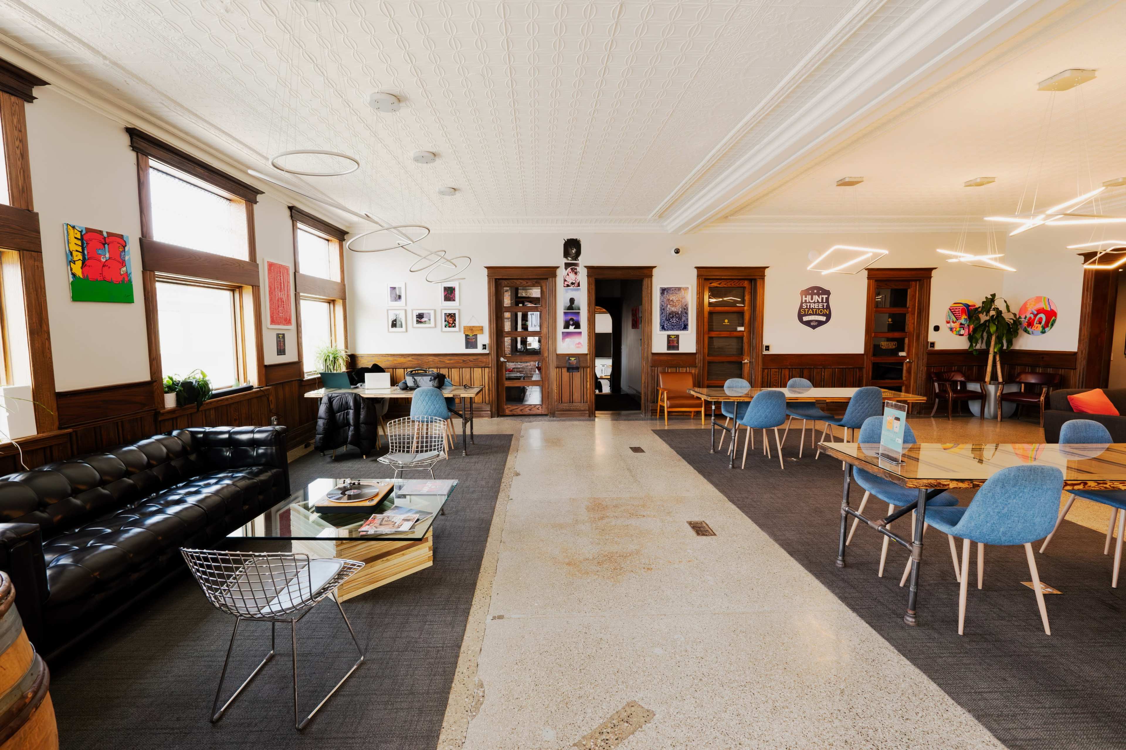 The image shows a spacious, well-lit interior of a modern lounge area featuring a mix of seating options, including a black leather couch, wire chairs, and wooden tables.