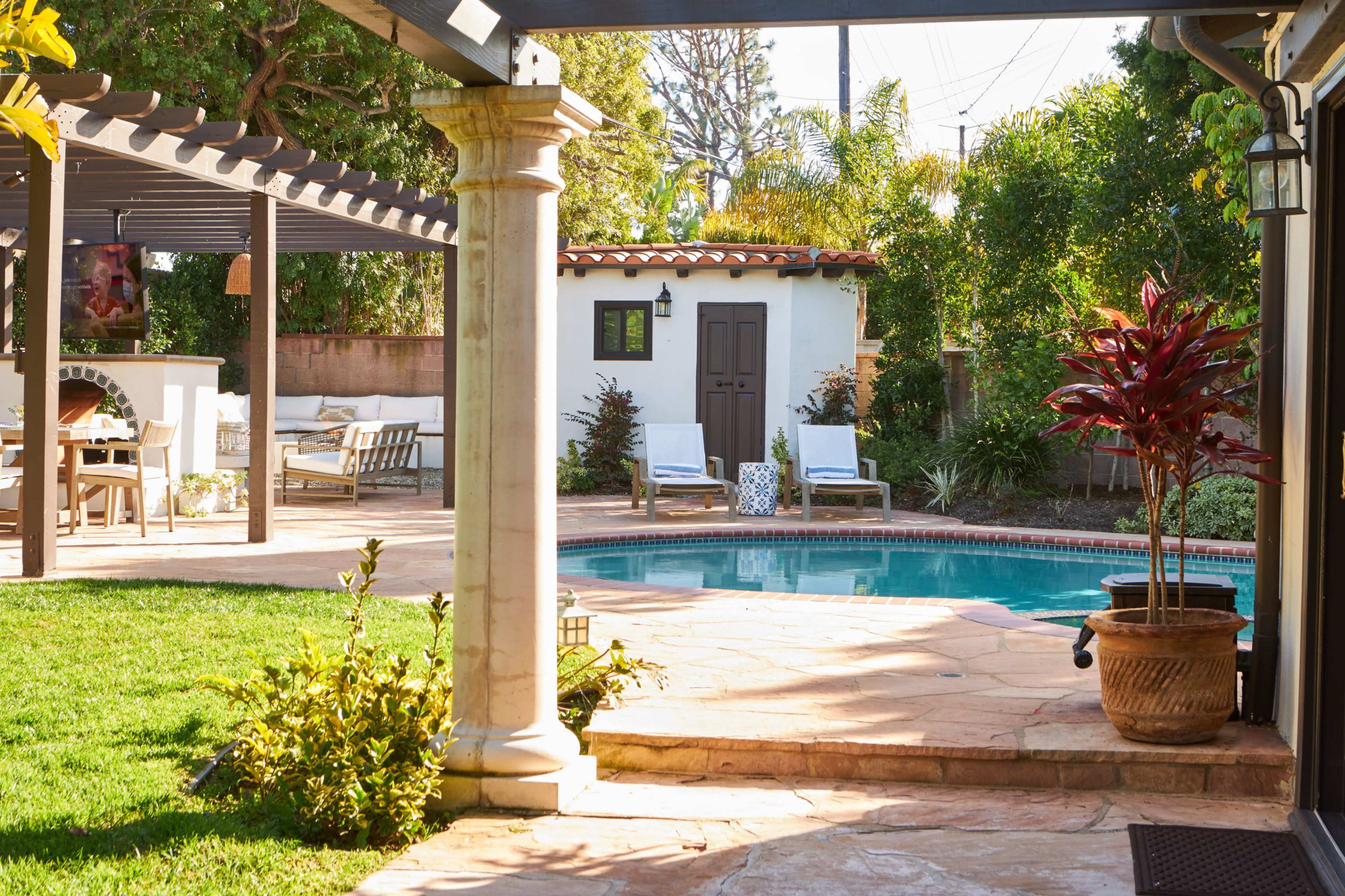 A stone patio with a swimming pool is surrounded by greenery and a small guest house in a residential backyard.