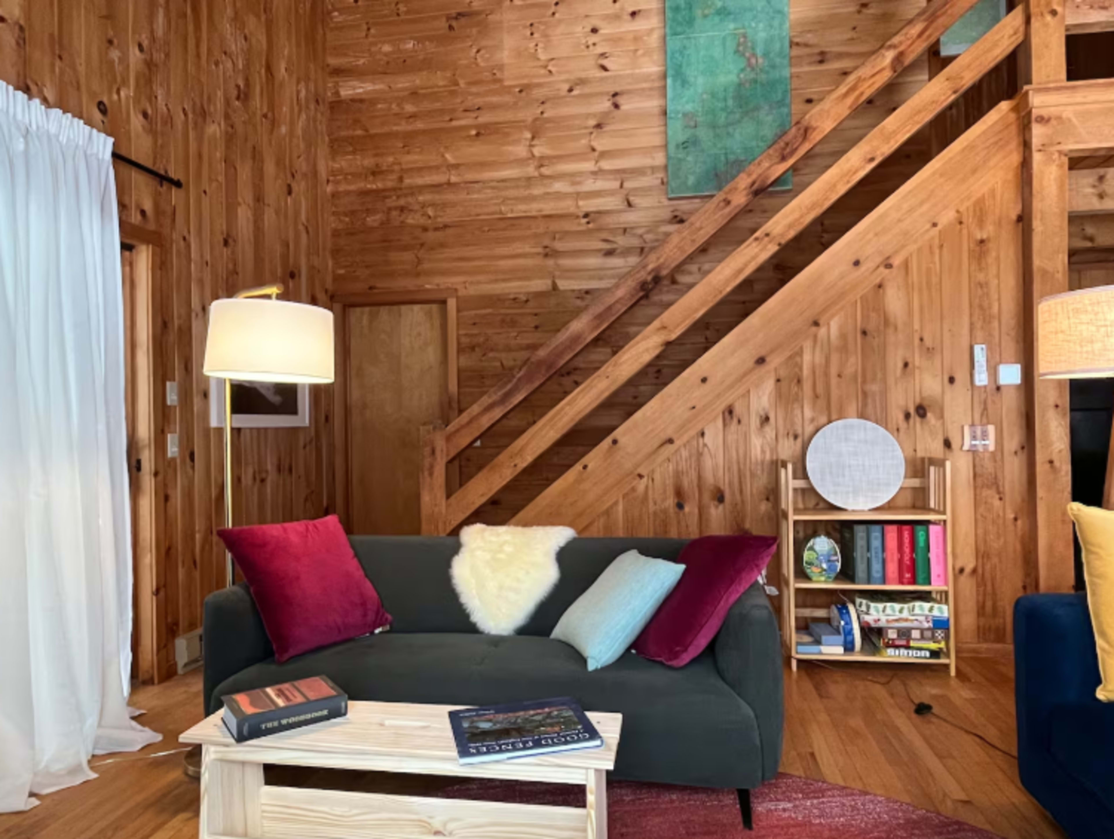 The image features a cozy wooden living room with a dark sofa adorned with colorful cushions, a small coffee table, and a staircase leading to an upper level.