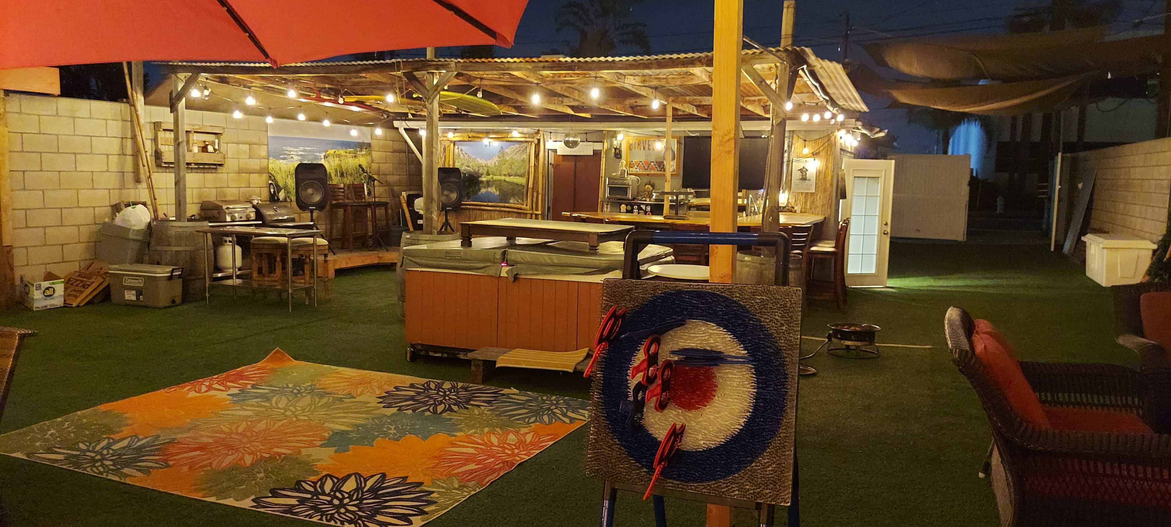 A backyard space at night with a dartboard, a colorful rug, various seating areas, and a covered patio illuminated by string lights.