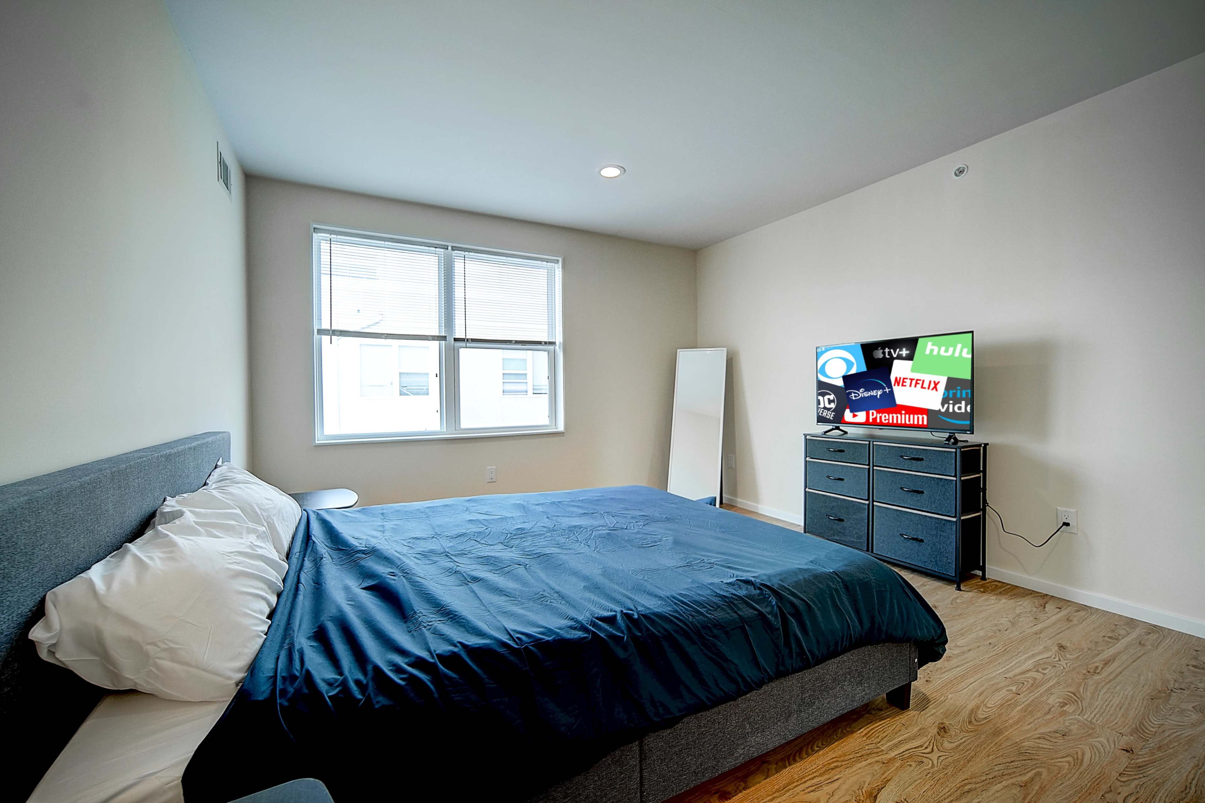 A bedroom with a large bed, a mirror, and a dresser featuring a television displaying various streaming service logos.
