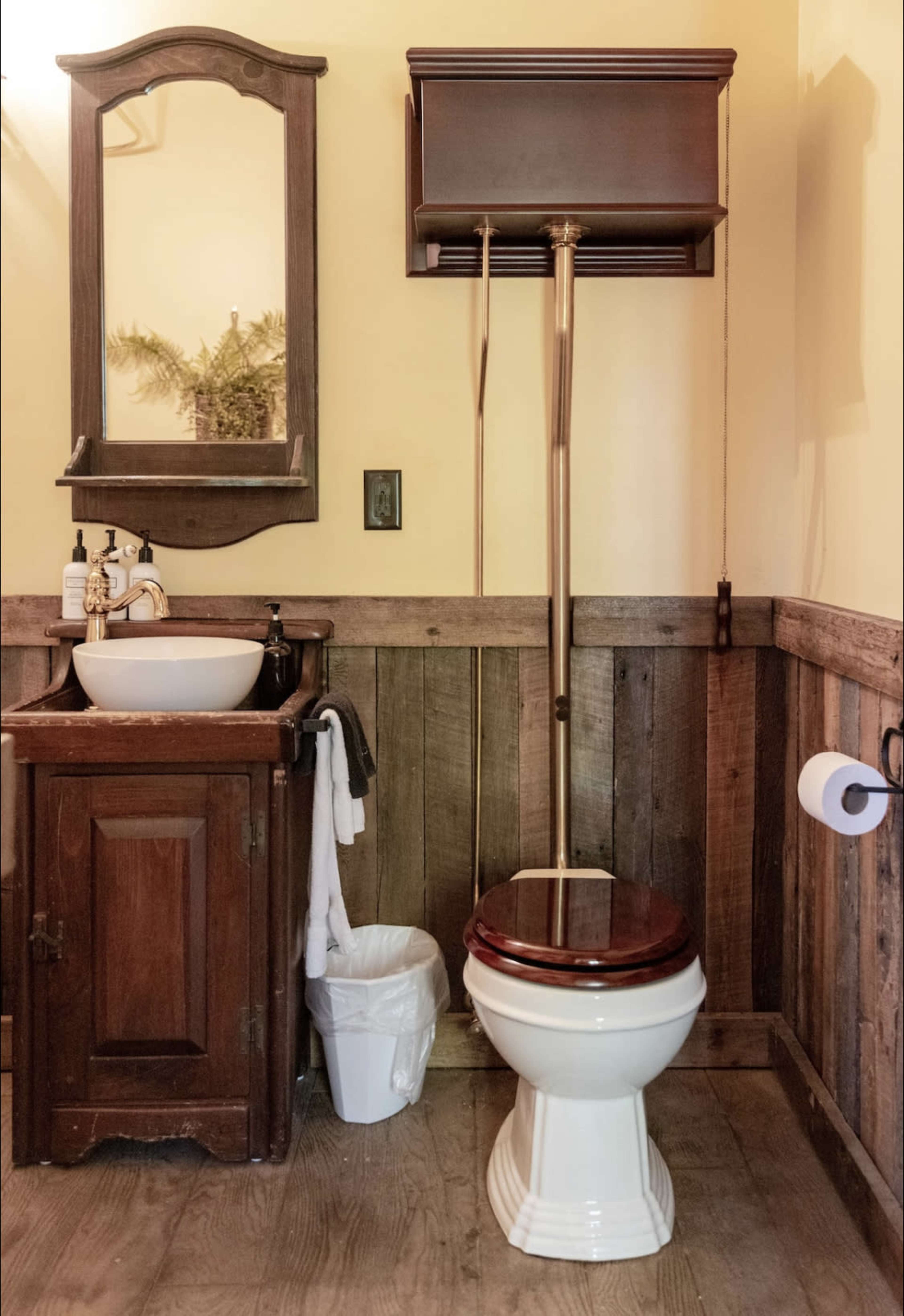 A bathroom featuring a wooden vanity with a sink, a toilet with a high tank, and a wall-mounted mirror.