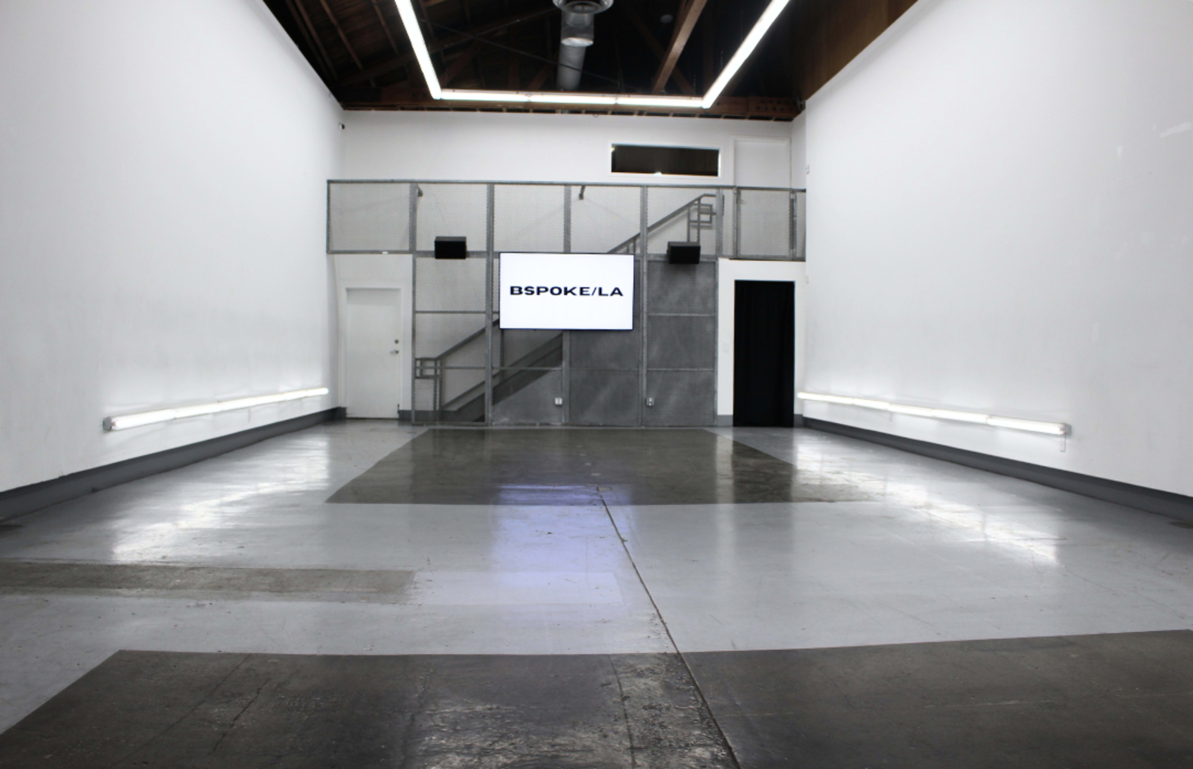 The image shows an empty, well-lit interior space with a large screen displaying "BESPOKE/LA" and a staircase in the background.