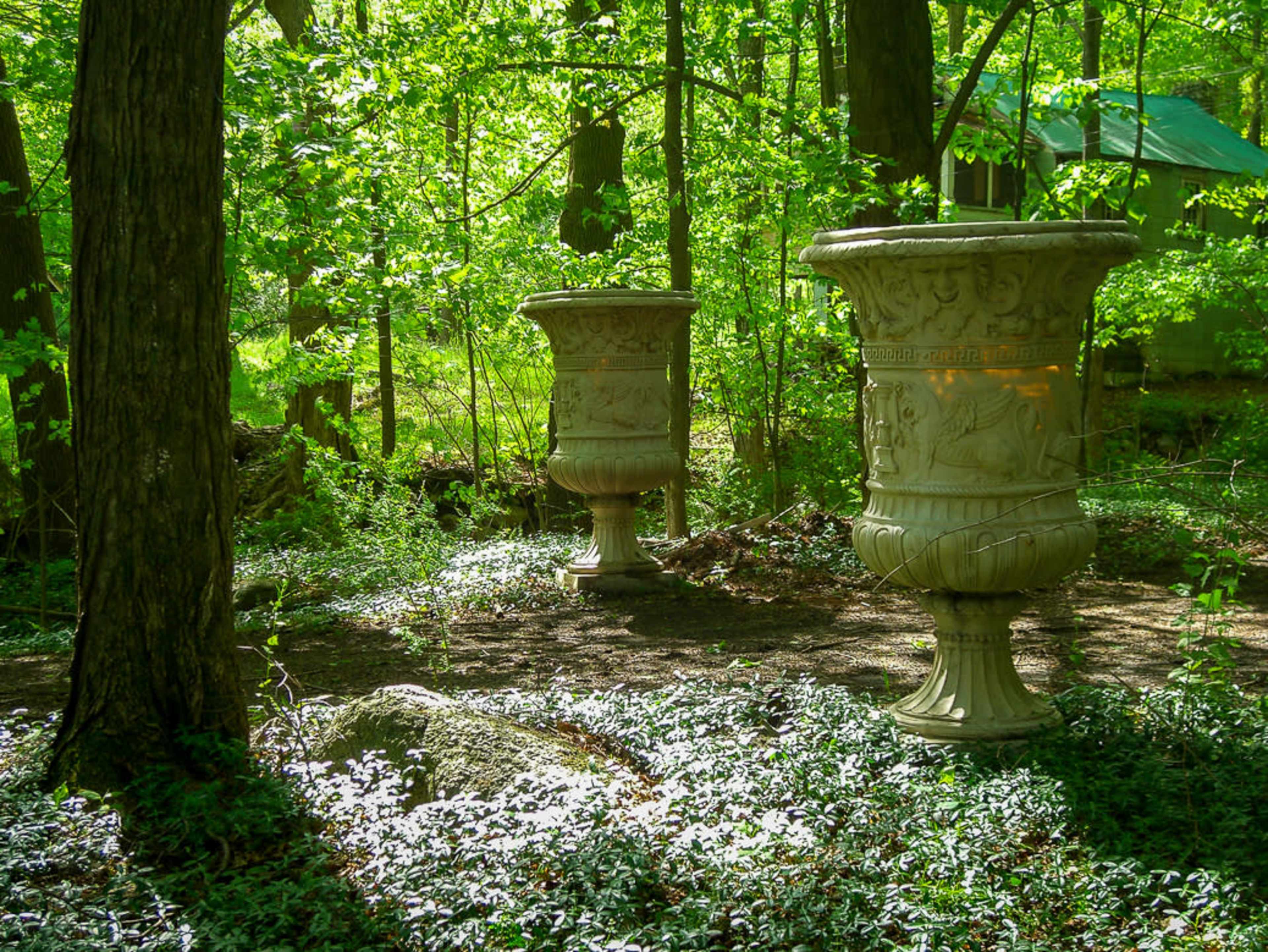 Two decorative garden urns stand amidst lush green foliage in a wooded area.