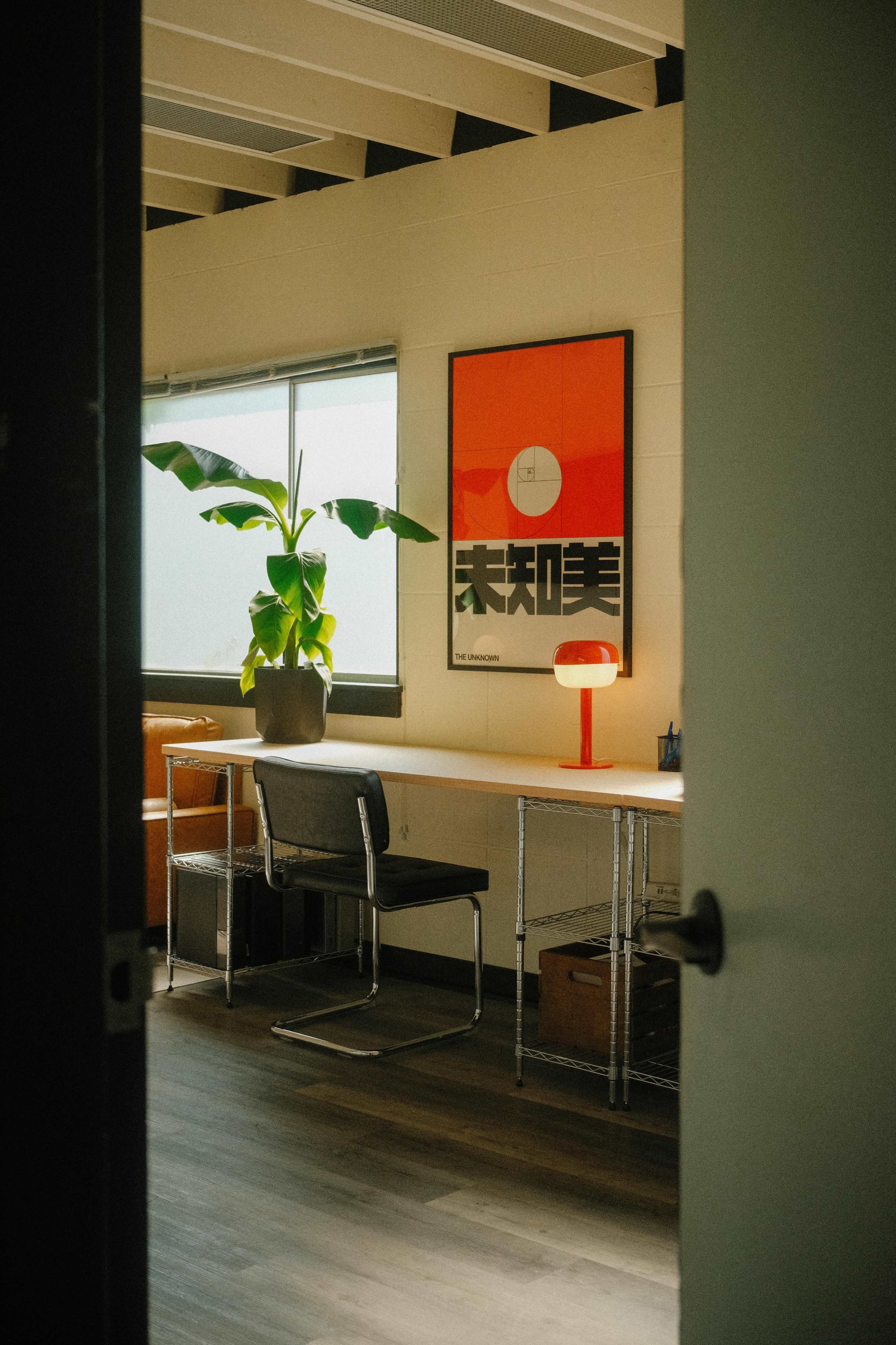 A modern workspace features a desk with a lamp and a chair, alongside a potted plant and a poster on the wall.