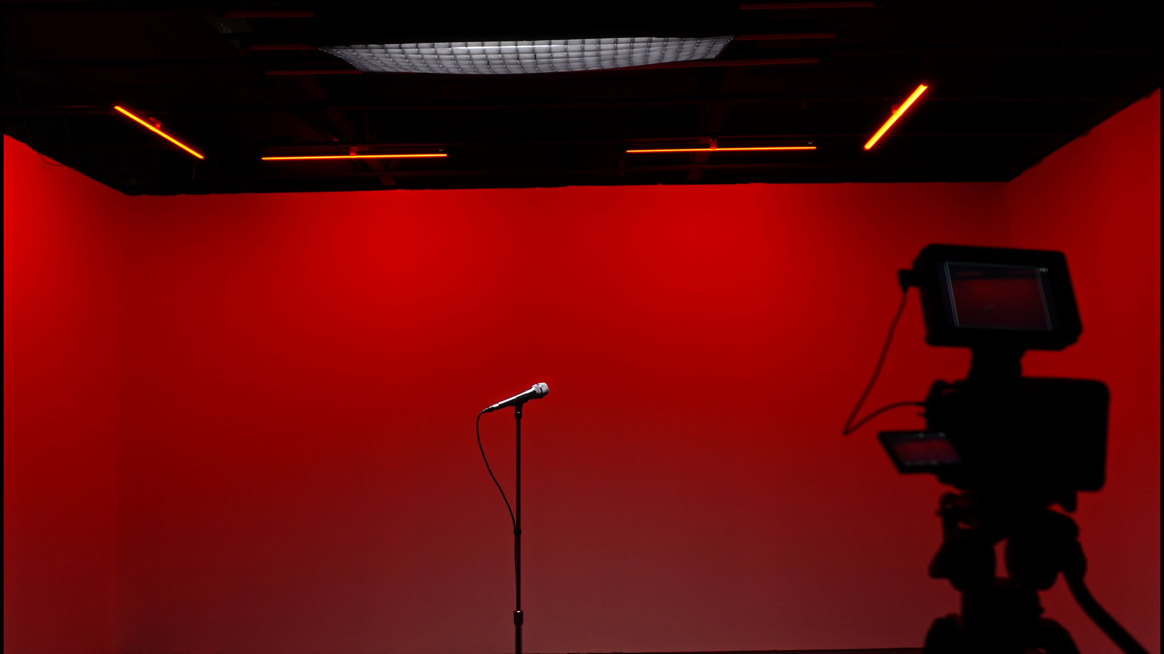 A microphone stands on a black stand in front of a solid red backdrop, with a camera silhouette capturing the scene.