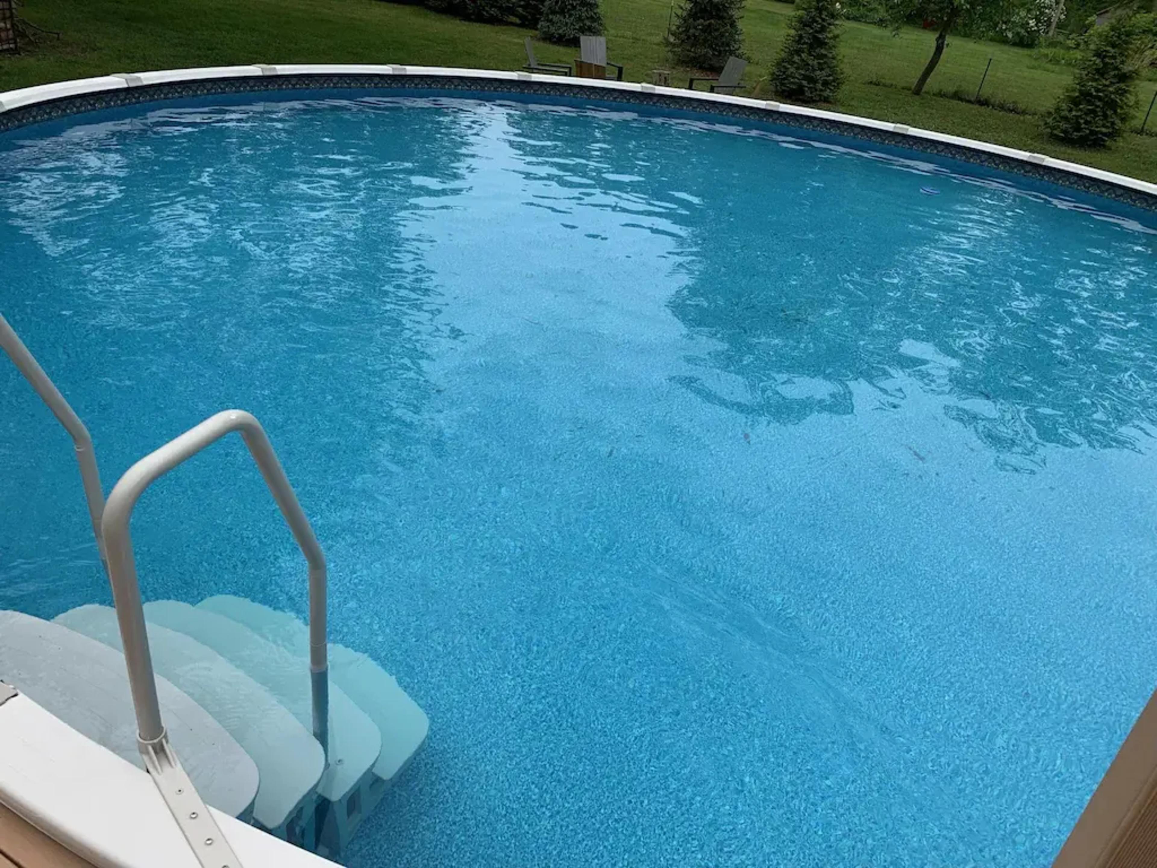 A round swimming pool with blue water and a set of white stairs leading into it, surrounded by a grassy area and trees in the background.