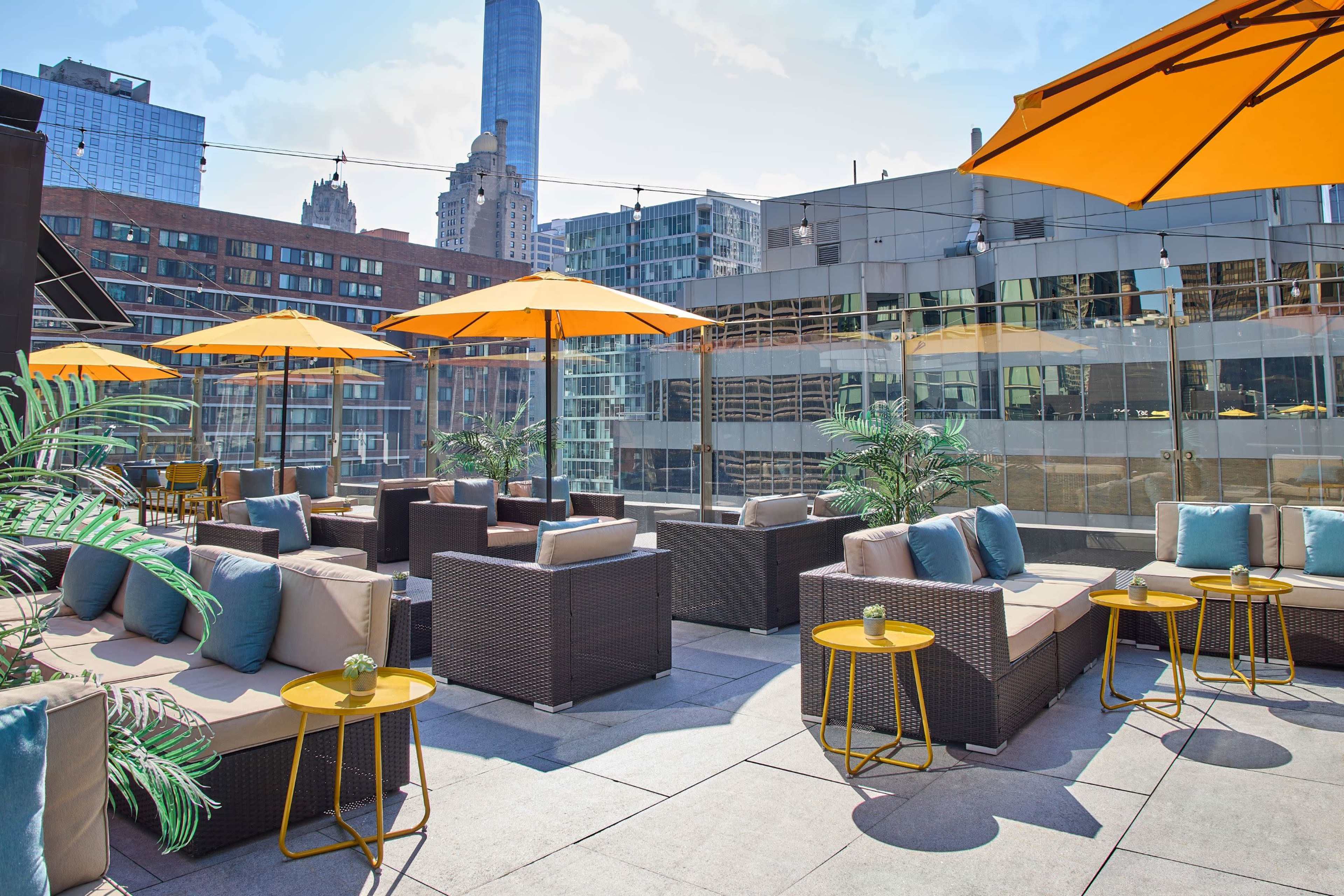 The image features a rooftop lounge area with modern furniture, vibrant orange umbrellas, and views of city buildings in the background.